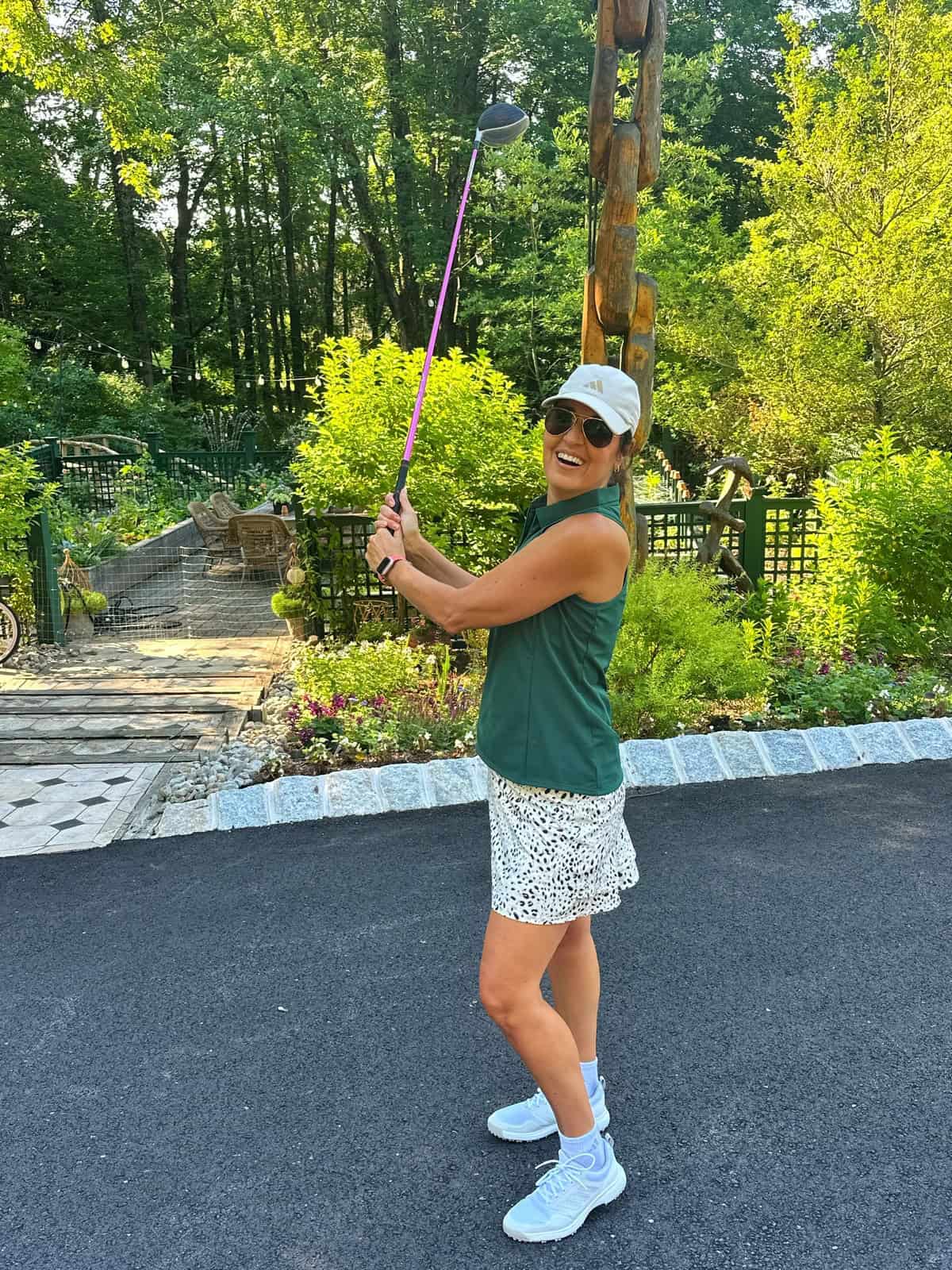 A smiling woman in a white cap, sunglasses, green sleeveless top, and white skirt holds a golf club outside on a paved path, surrounded by greenery and gardens on a sunny day.
