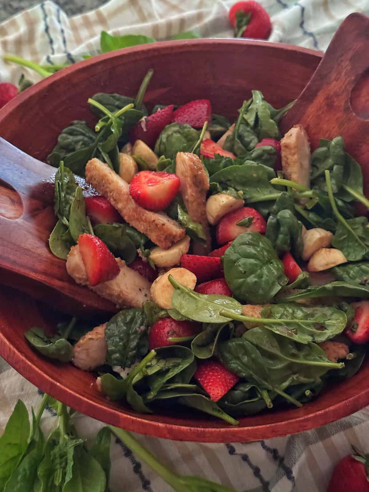 A wooden bowl filled with a fresh spinach salad with strawberries, grilled chicken strips, and cashews, with wooden salad tongs resting in the bowl.