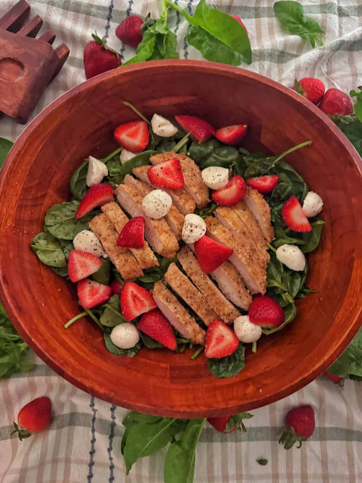 A wooden bowl filled with spinach salad with strawberries, sliced breaded chicken, and mozzarella balls sits on a light plaid cloth, with extra spinach and halved strawberries scattered nearby.