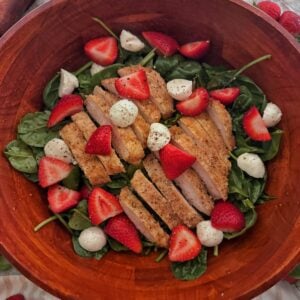 A wooden bowl filled with spinach salad with strawberries, sliced breaded chicken, mozzarella balls, and black pepper, garnished to perfection. Extra spinach leaves and strawberries are scattered around for a fresh touch.