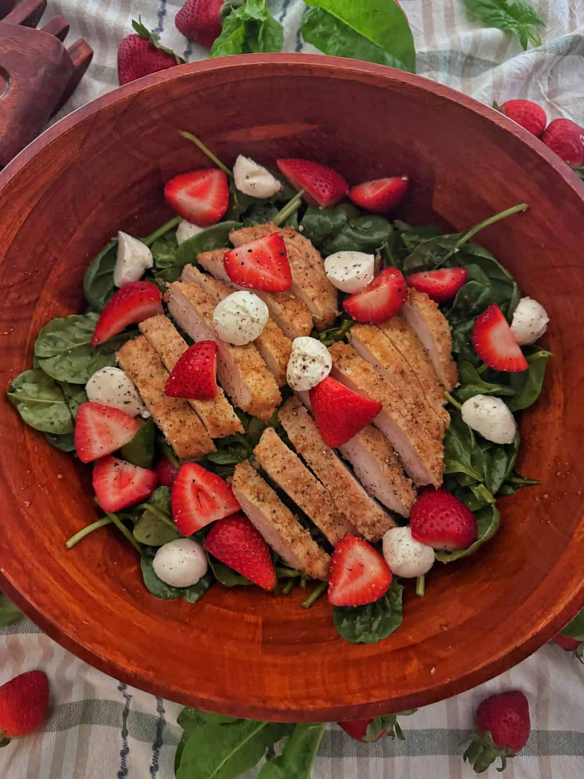 A wooden bowl filled with spinach salad with strawberries, sliced breaded chicken, mozzarella balls, and black pepper, garnished to perfection. Extra spinach leaves and strawberries are scattered around for a fresh touch.