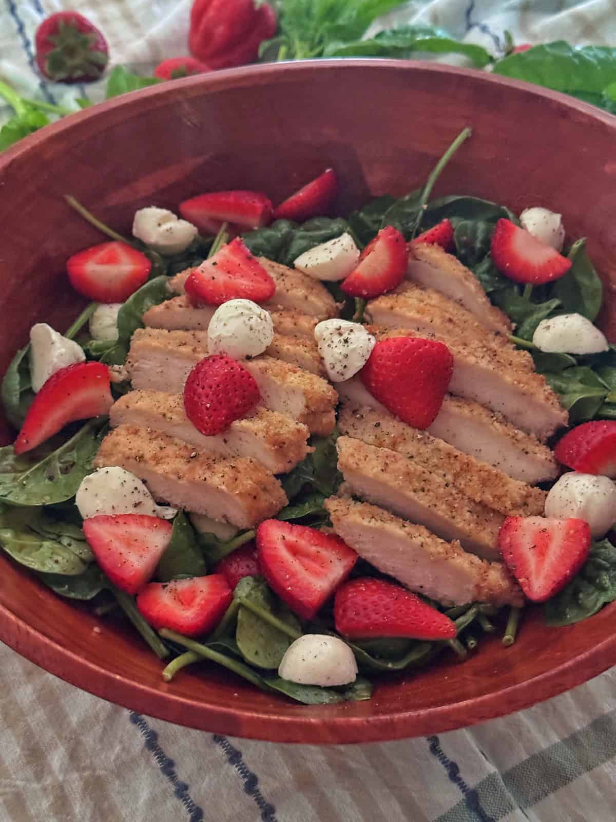 A wooden bowl filled with a vibrant spinach salad with strawberries, sliced breaded chicken, and small mozzarella balls, garnished with cracked black pepper.