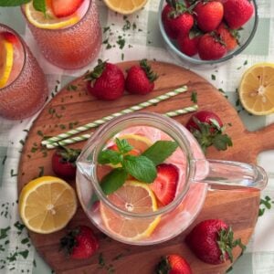 A pitcher of strawberry lemonade with lemon and strawberry slices, garnished with mint, on a round wooden board. Surrounded by fresh strawberries, lemon halves, two glasses of lemonade, and green-striped straws.