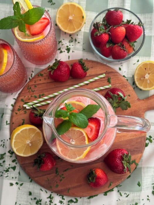 A pitcher of strawberry lemonade with lemon and strawberry slices, garnished with mint, on a round wooden board. Surrounded by fresh strawberries, lemon halves, two glasses of lemonade, and green-striped straws.