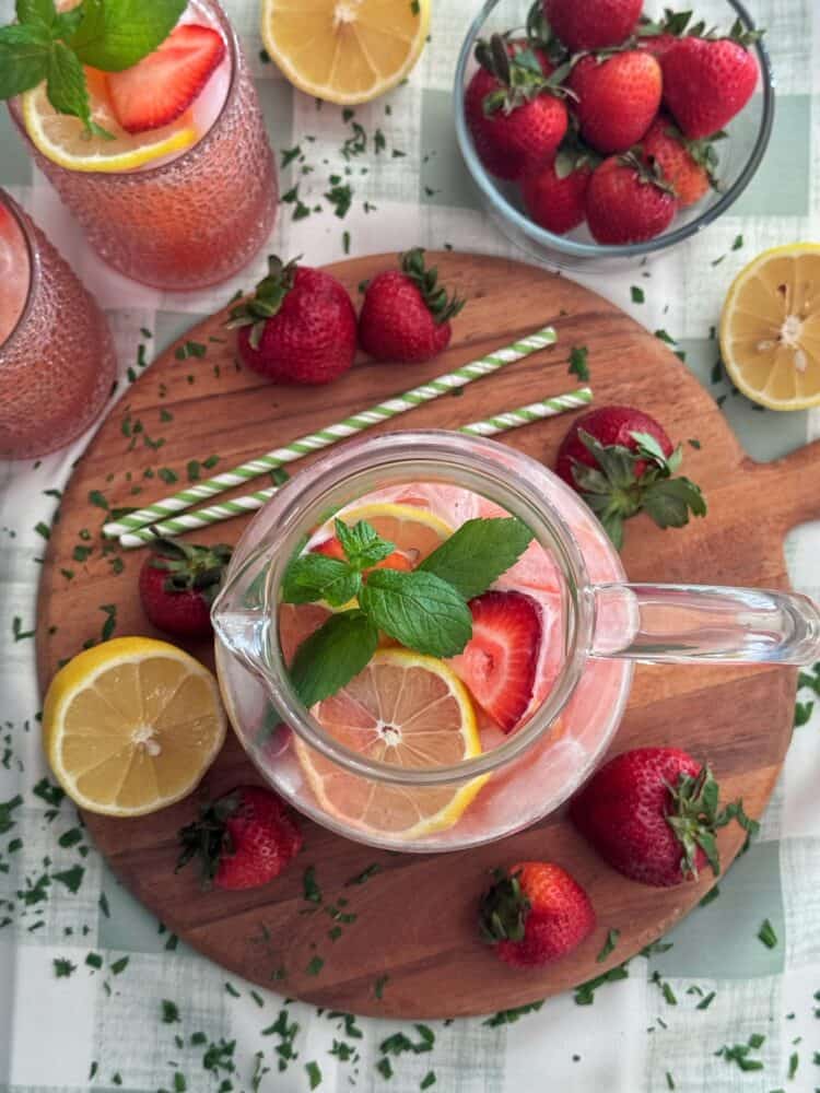 A pitcher of strawberry lemon lemonade with mint sits on a wooden board surrounded by fresh strawberries, lemon halves, two glasses of the drink, and green striped straws on a light checkered tablecloth.