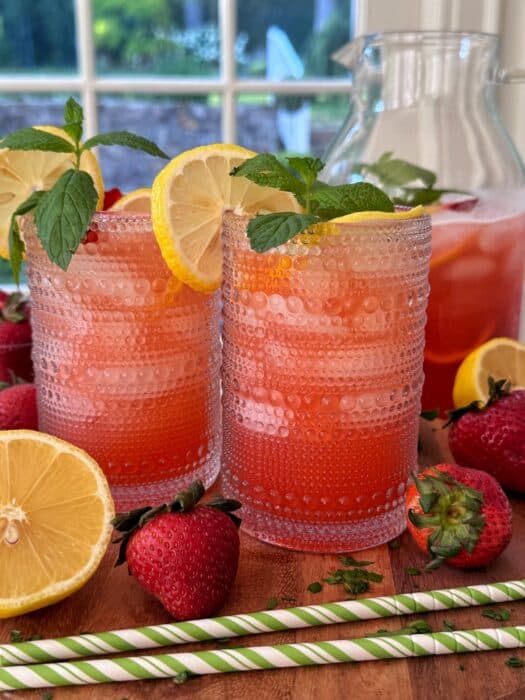 Two textured glasses filled with iced strawberry lemonade, garnished with lemon slices and mint leaves, sit on a wooden table with fresh strawberries and lemon halves, next to a glass pitcher and striped paper straws.