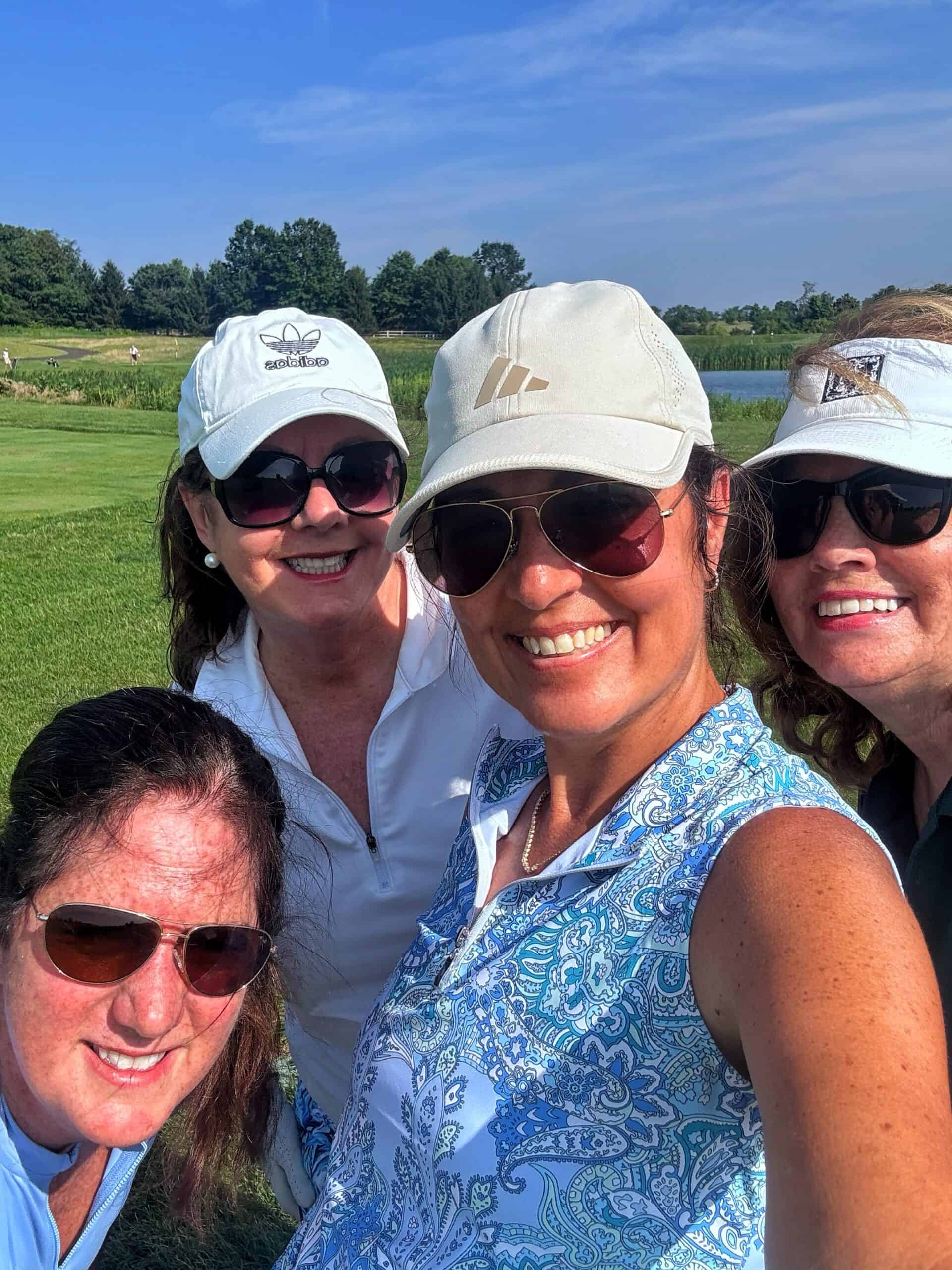 Four women (one is stacy ling) in sunglasses and hats smile for a selfie on a sunny golf course, with green grass, trees, and a pond visible in the background.