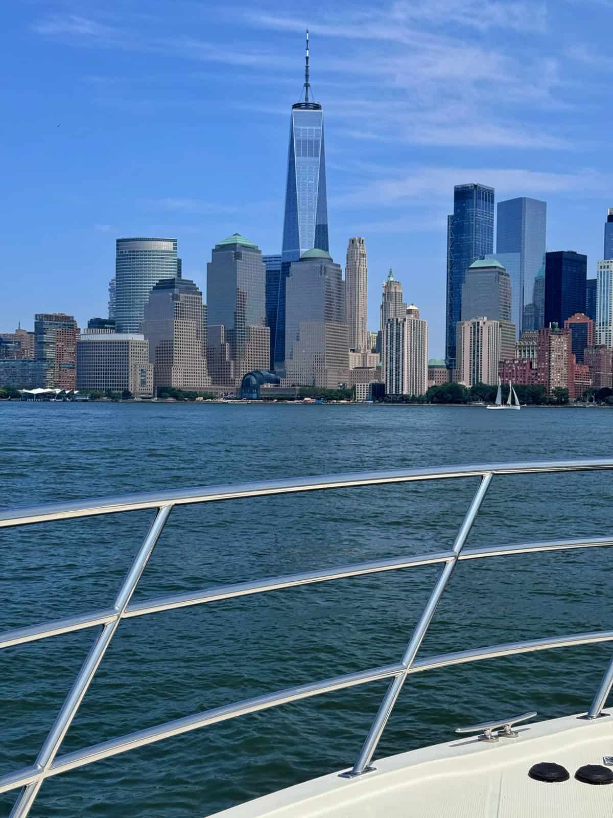 View of the New York City skyline, including One World Trade Center, seen from a boat on the water with part of the boat’s railing and deck visible in the foreground.