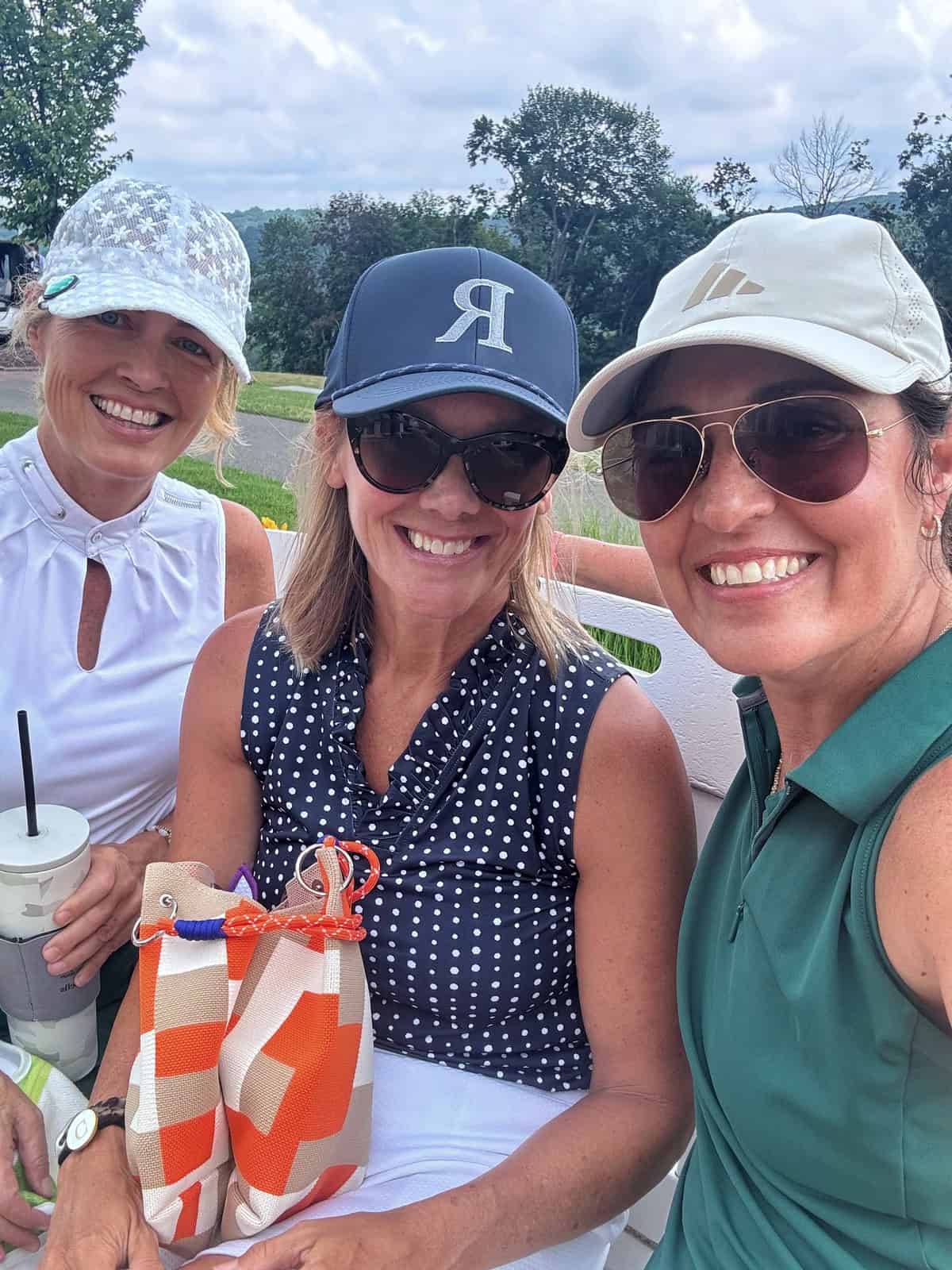 (Stacy Ling and friends) Three women in golf attire smile for a selfie outdoors. One holds a drink, another holds a colorful pouch, and trees and clouds are visible in the background.