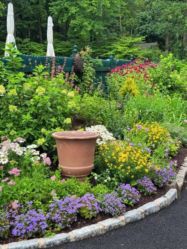 A colorful garden bed features blooming flowers in shades of purple, yellow, white, and pink around a large empty terracotta pot, bordered by a stone edge and surrounded by lush greenery.