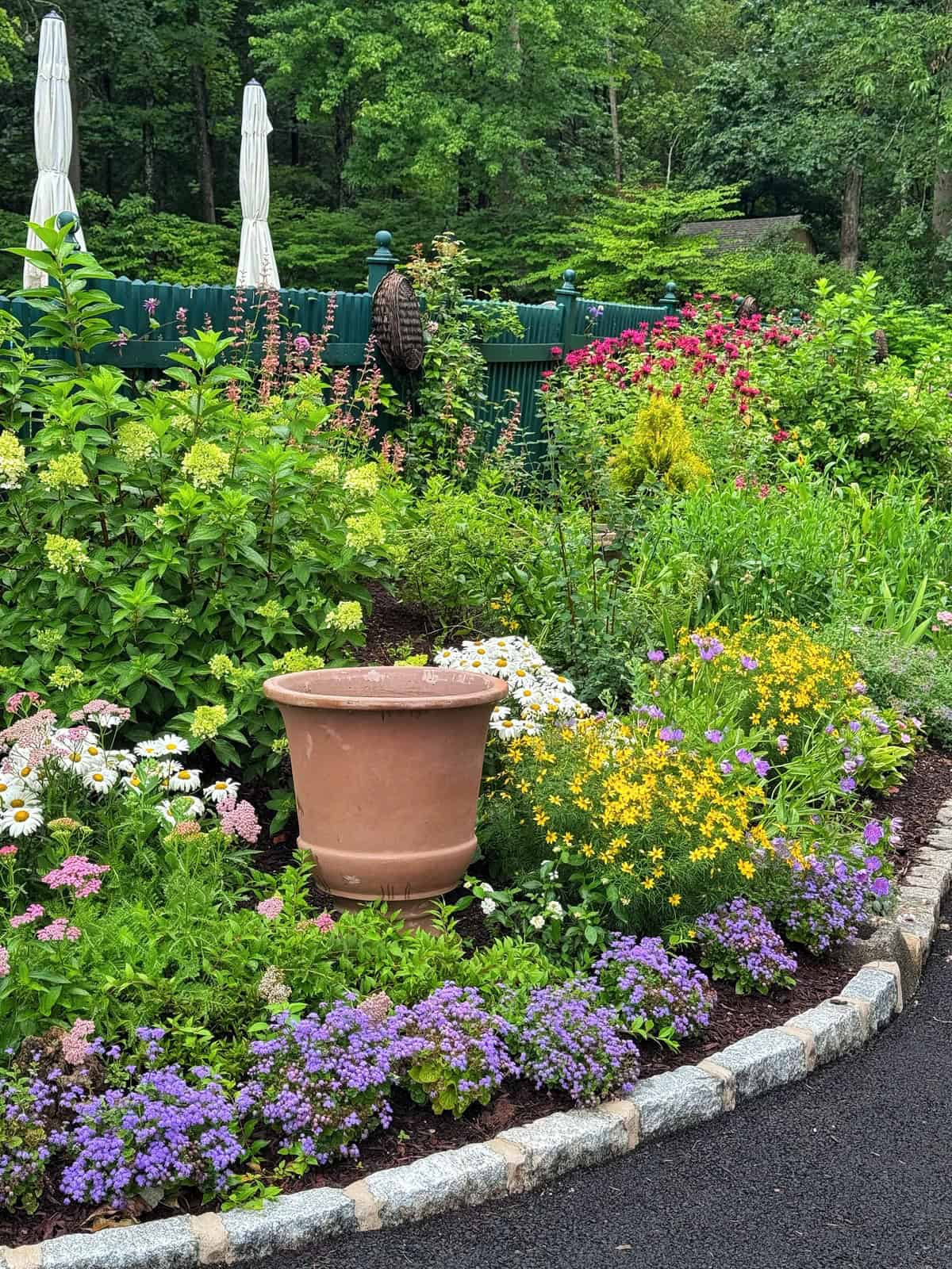 A colorful garden bed features blooming flowers in shades of purple, yellow, white, and pink around a large empty terracotta pot, bordered by a stone edge and surrounded by lush greenery.