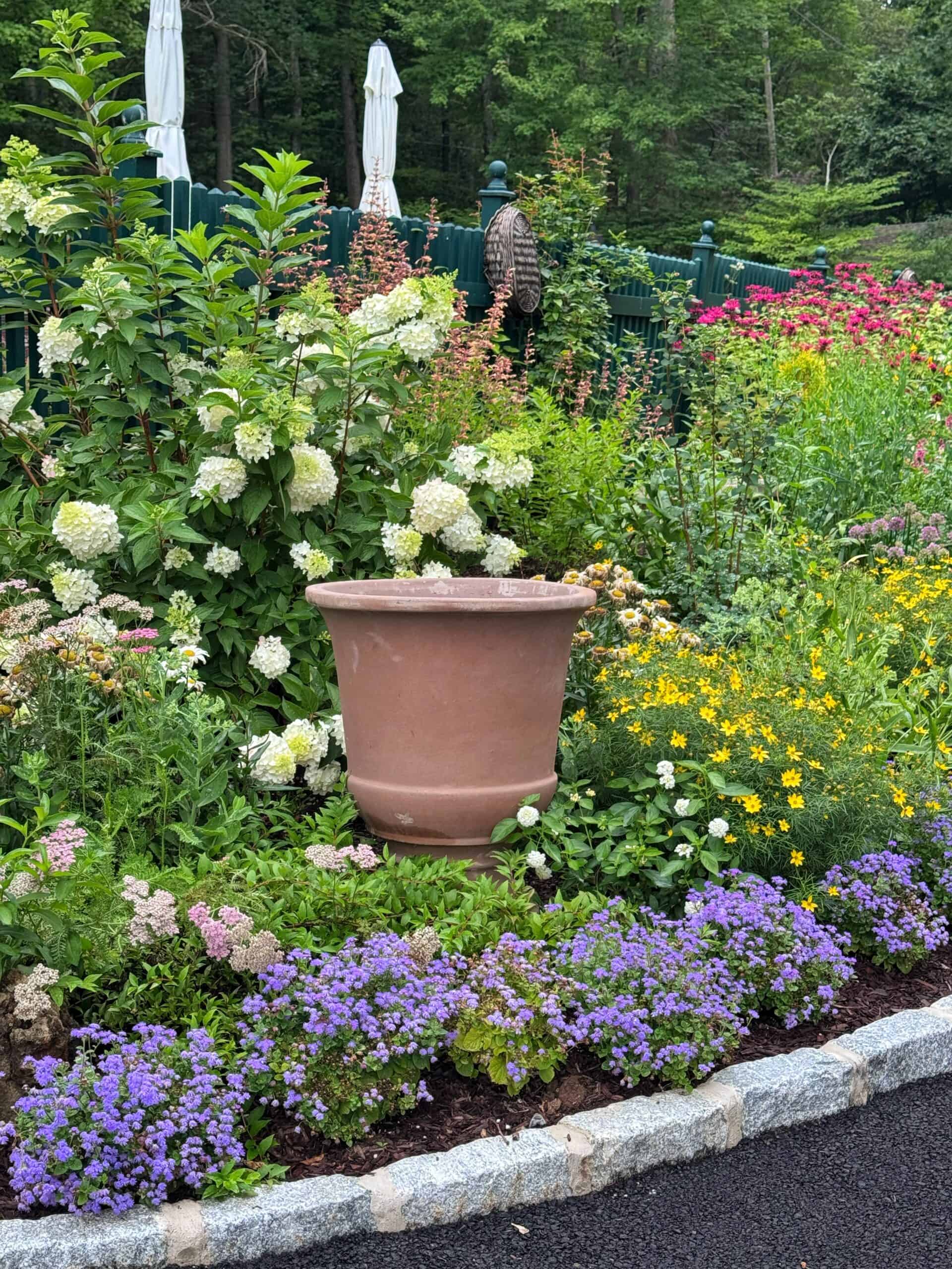 A large empty flower pot sits among colorful blooming flowers and green plants in a garden, with a stone edge and a person standing in the background.