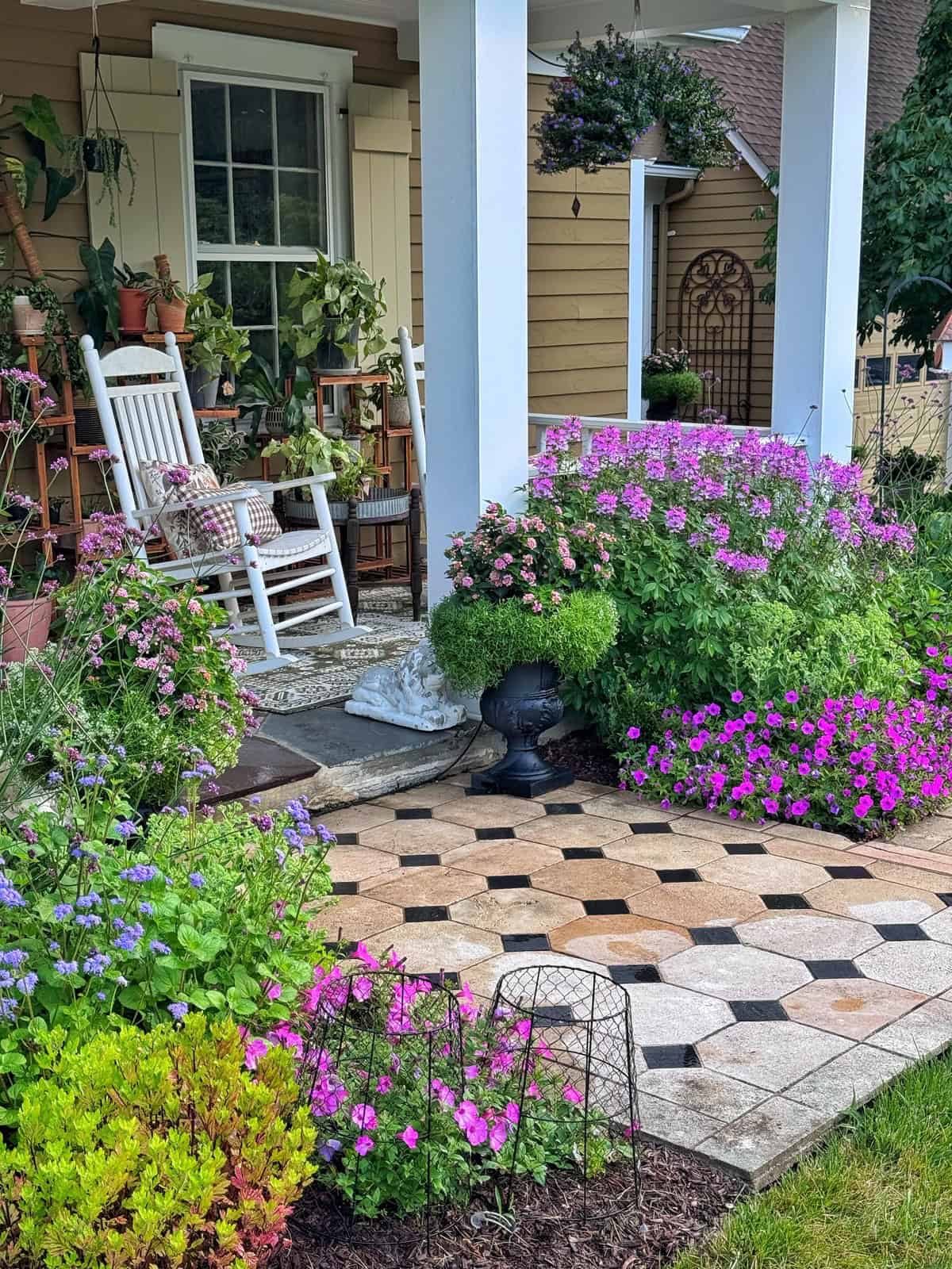A cozy porch with rocking chairs, potted plants, and colorful flowers lining a patterned stone walkway. Lush greenery and blooming pink, purple, and blue flowers create a vibrant, welcoming garden scene.