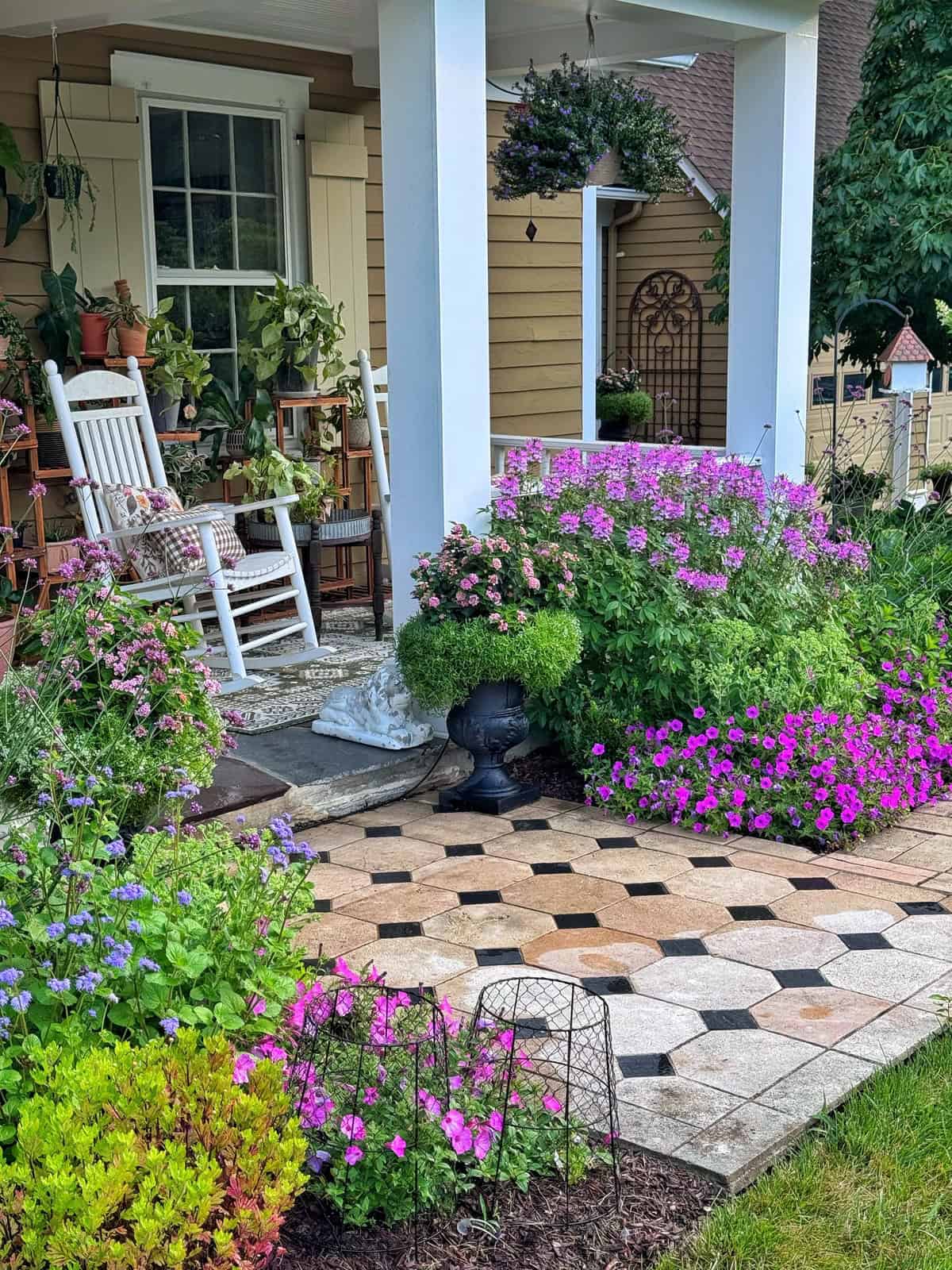 A cozy porch with rocking chairs, potted plants, and pillows overlooks a garden path lined with blooming pink, purple, and blue flowers and lush greenery. A hanging basket and garden gate are visible in the background.