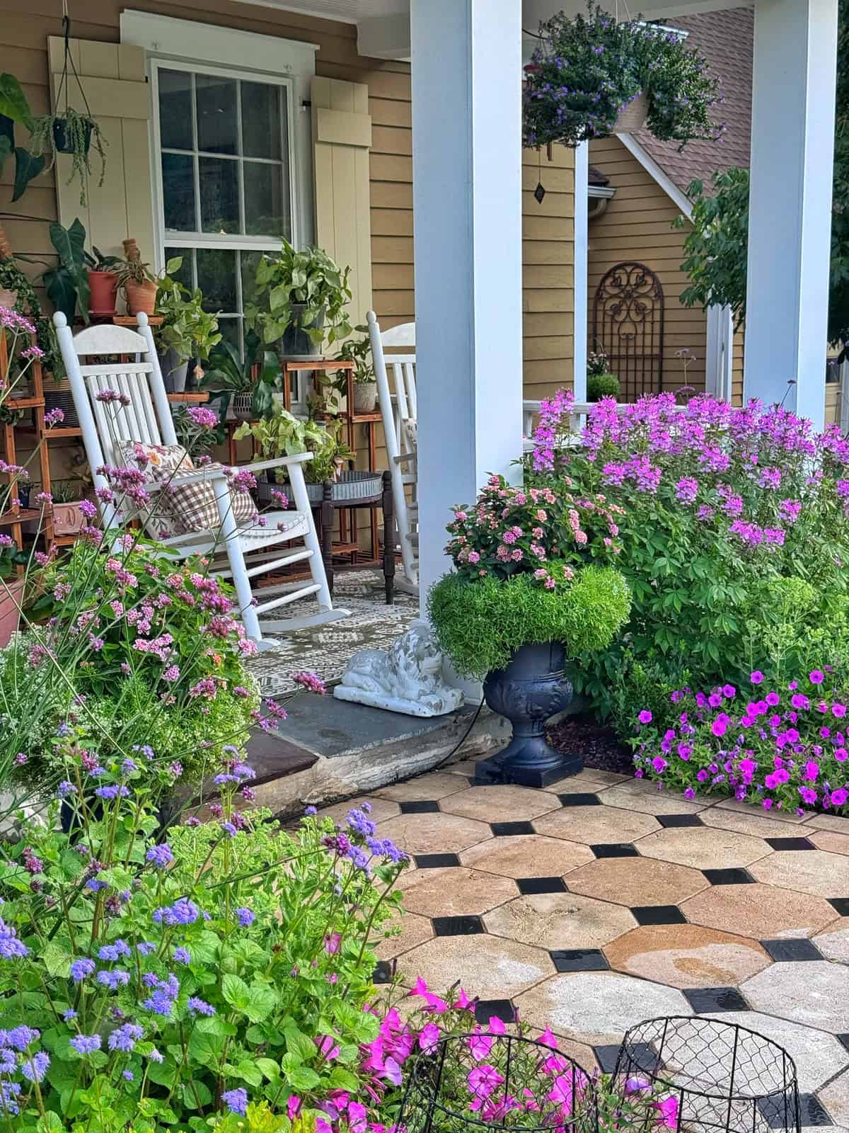 A cozy porch with white rocking chairs, potted plants, and pillows overlooks a vibrant garden filled with blooming purple and pink flowers. The scene feels inviting and peaceful.