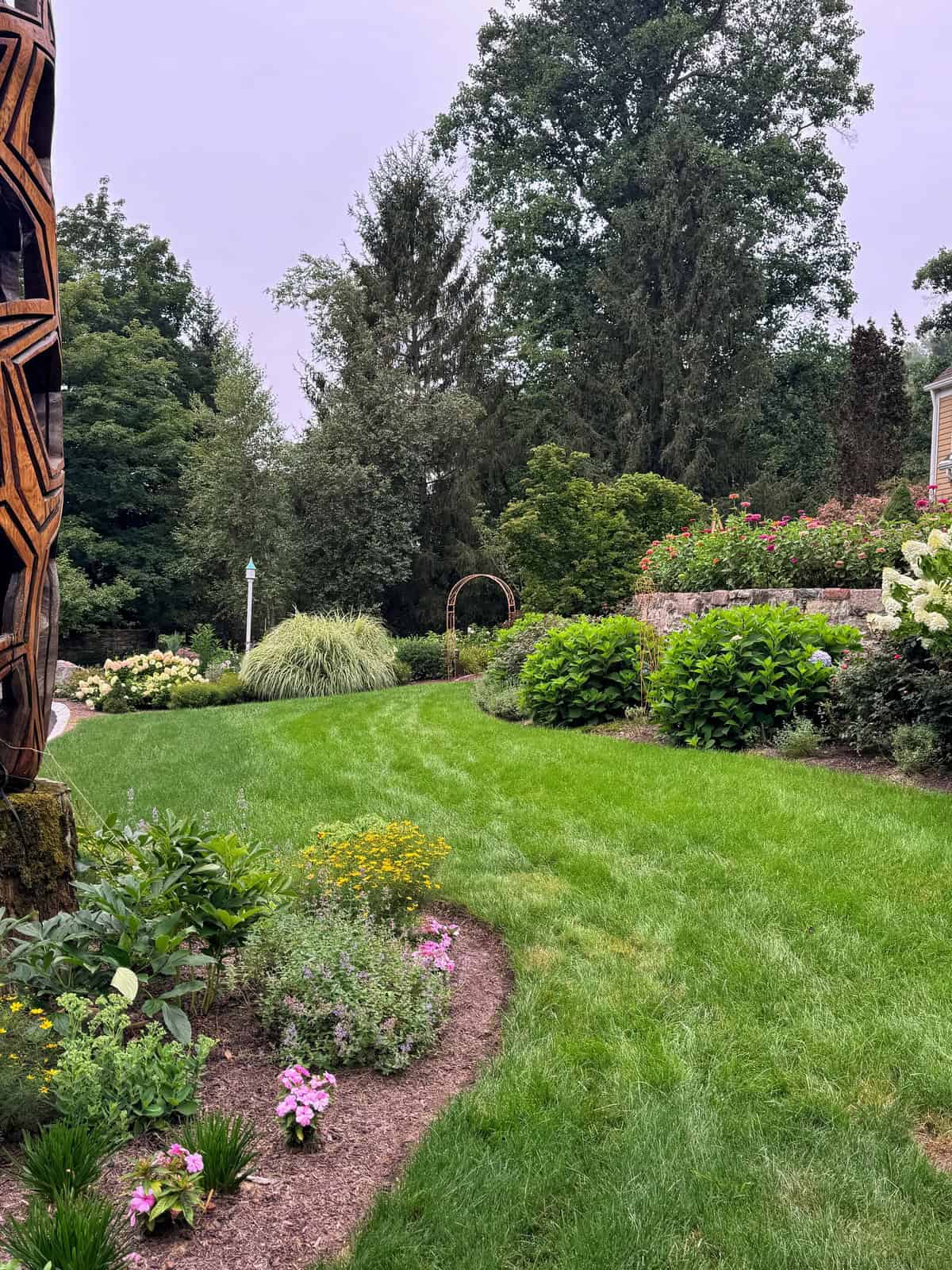 A lush green lawn bordered by colorful flower beds, tall trees, and shrubs. A circular wooden arch and a wooden sculpture are visible, with a house partially seen on the right.
