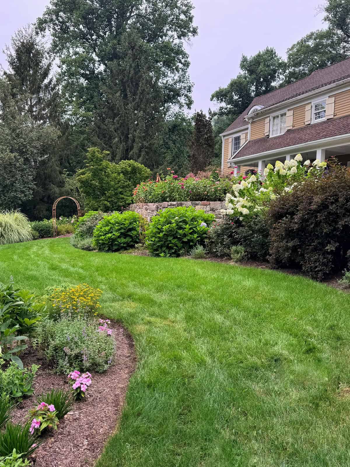 A well-maintained garden with green grass, flowering plants, shrubs, and a stone retaining wall next to a tan house. A wooden archway stands at the back among trees.