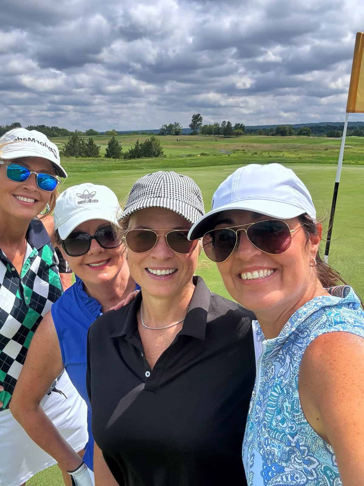 Four women in golf attire smile for a selfie on a golf course under a cloudy sky. The green field and a flagstick are visible in the background.