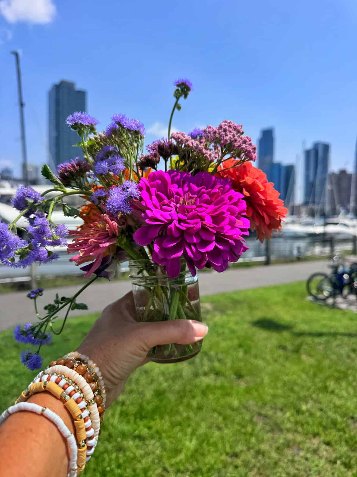 A hand wearing beaded bracelets holds a small glass jar with vibrant flowers. In the background are boats in a marina, green grass, and tall city buildings under a clear blue sky.