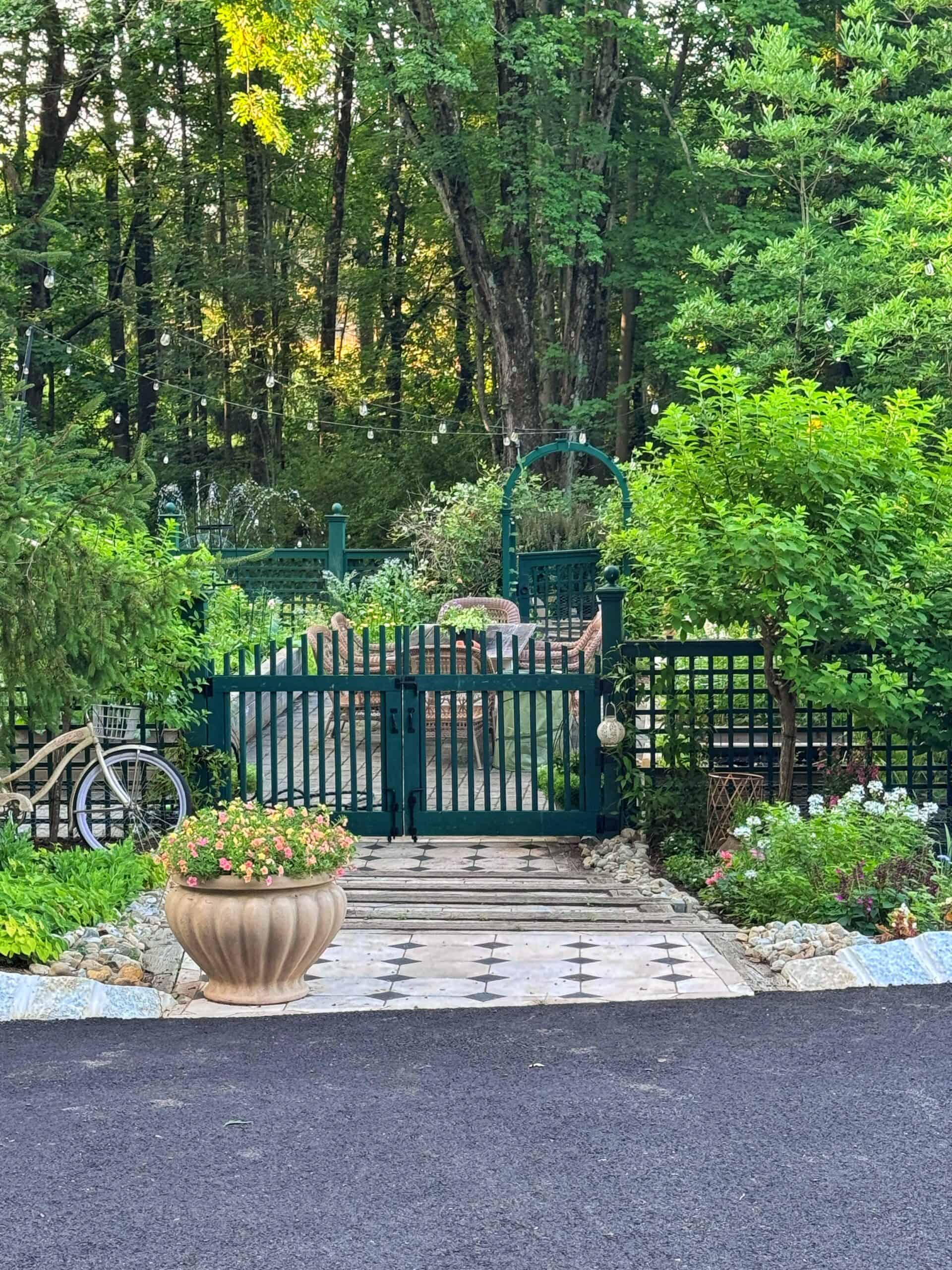 A lush garden with green fencing, a decorative archway, and string lights hangs above. A potted plant sits by the entrance, a vintage bike leans on the fence, and dense trees fill the background.