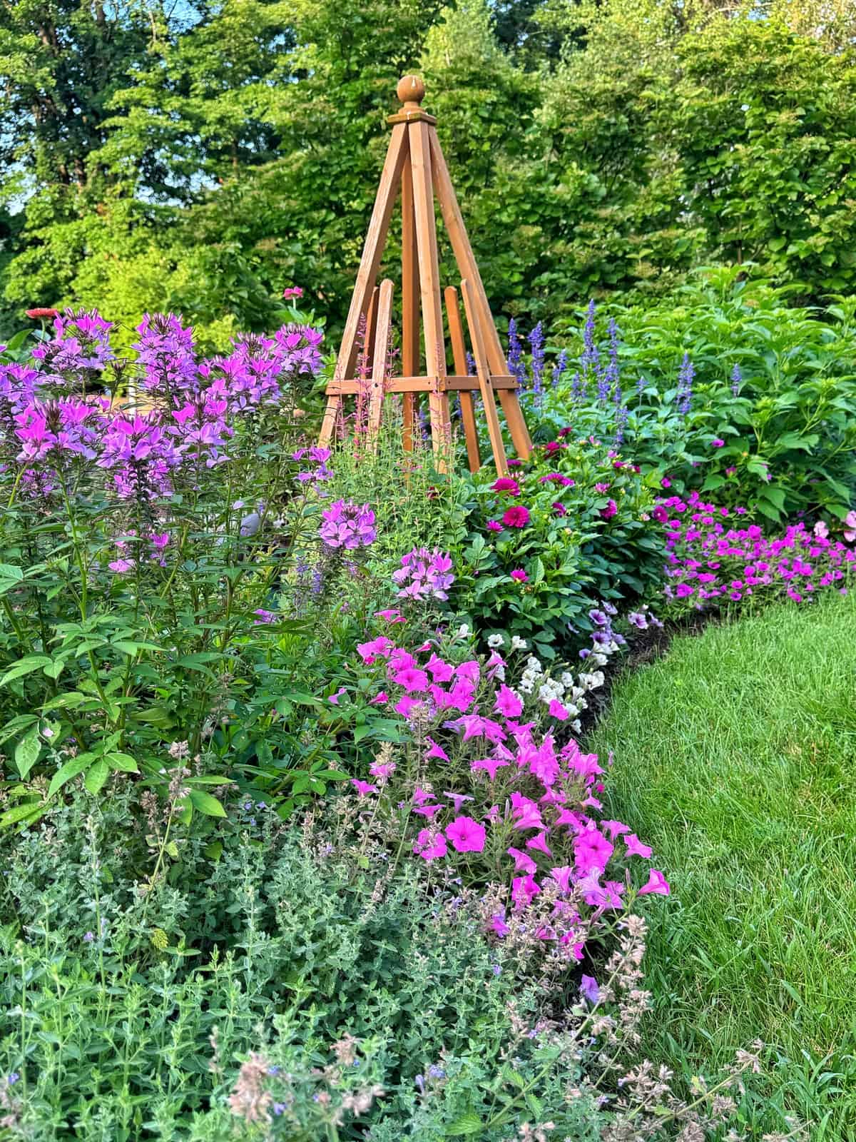 A vibrant garden bed with pink, purple, and blue flowers surrounds a wooden trellis, set against a lush, green background of trees and grass.