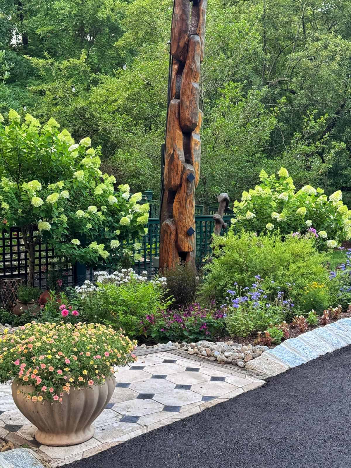 A lush garden with blooming flowers and green shrubs surrounds a tall wooden sculpture. A large stone planter with colorful flowers sits on a tiled patio next to a paved walkway. Trees fill the background.
