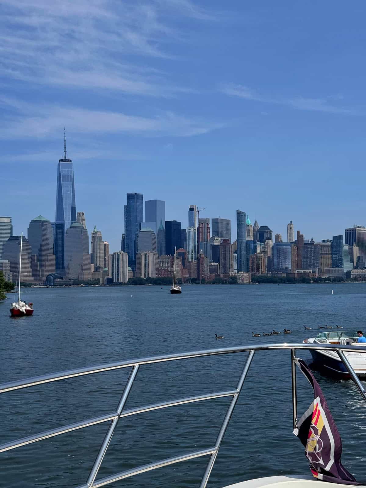 View of the New York City skyline from a boat on the water, with the One World Trade Center visible and a flag on the boat's railing in the foreground. Sailboats float nearby under a blue sky.