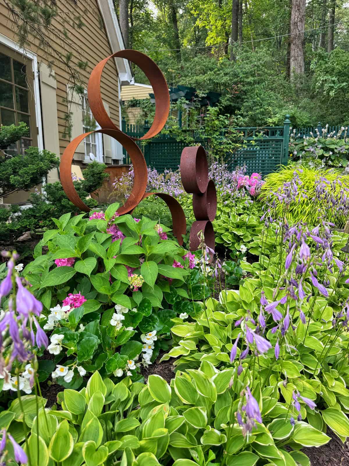A garden features green plants and purple flowers, with two rust-colored metal sculptures made of stacked circles. A tan house and trees are visible in the background.