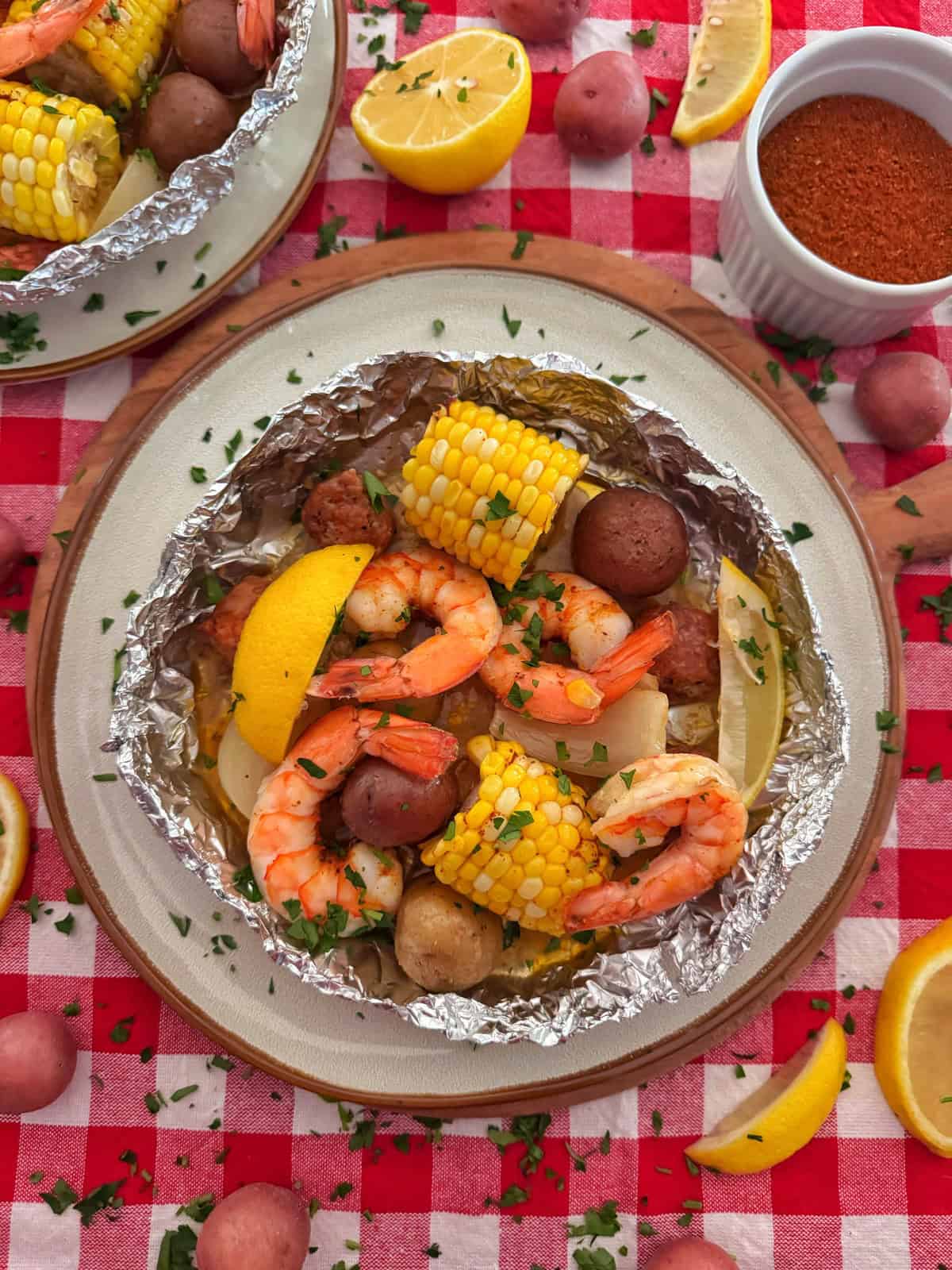 A shrimp boil foil packet on a plate holds shrimp, corn on the cob, red potatoes, sausage, and lemon wedges, garnished with herbs. The plate sits on a red checkered tablecloth with more potatoes, lemon slices, and a ramekin of seasoning nearby.