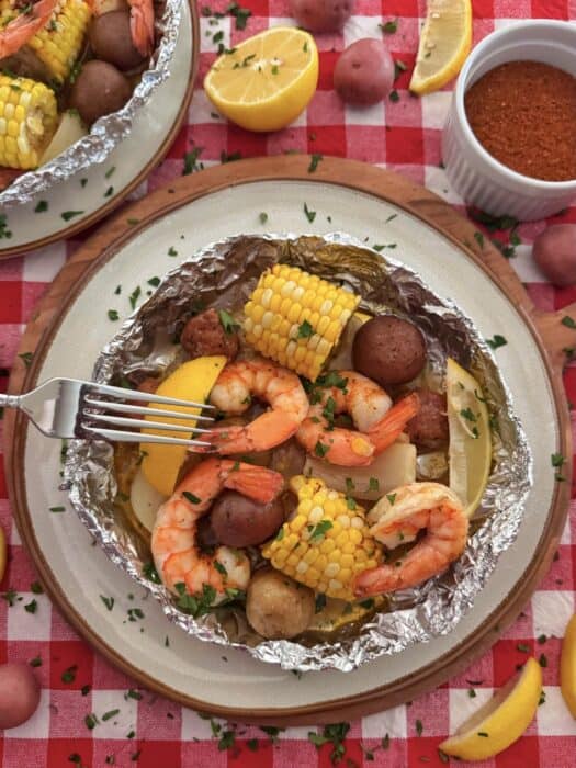 A plate with shrimp, corn on the cob, red potatoes, and sausage in foil, garnished with herbs. A fork, lemon wedges, seasoning, and more food are on a red checkered tablecloth.
