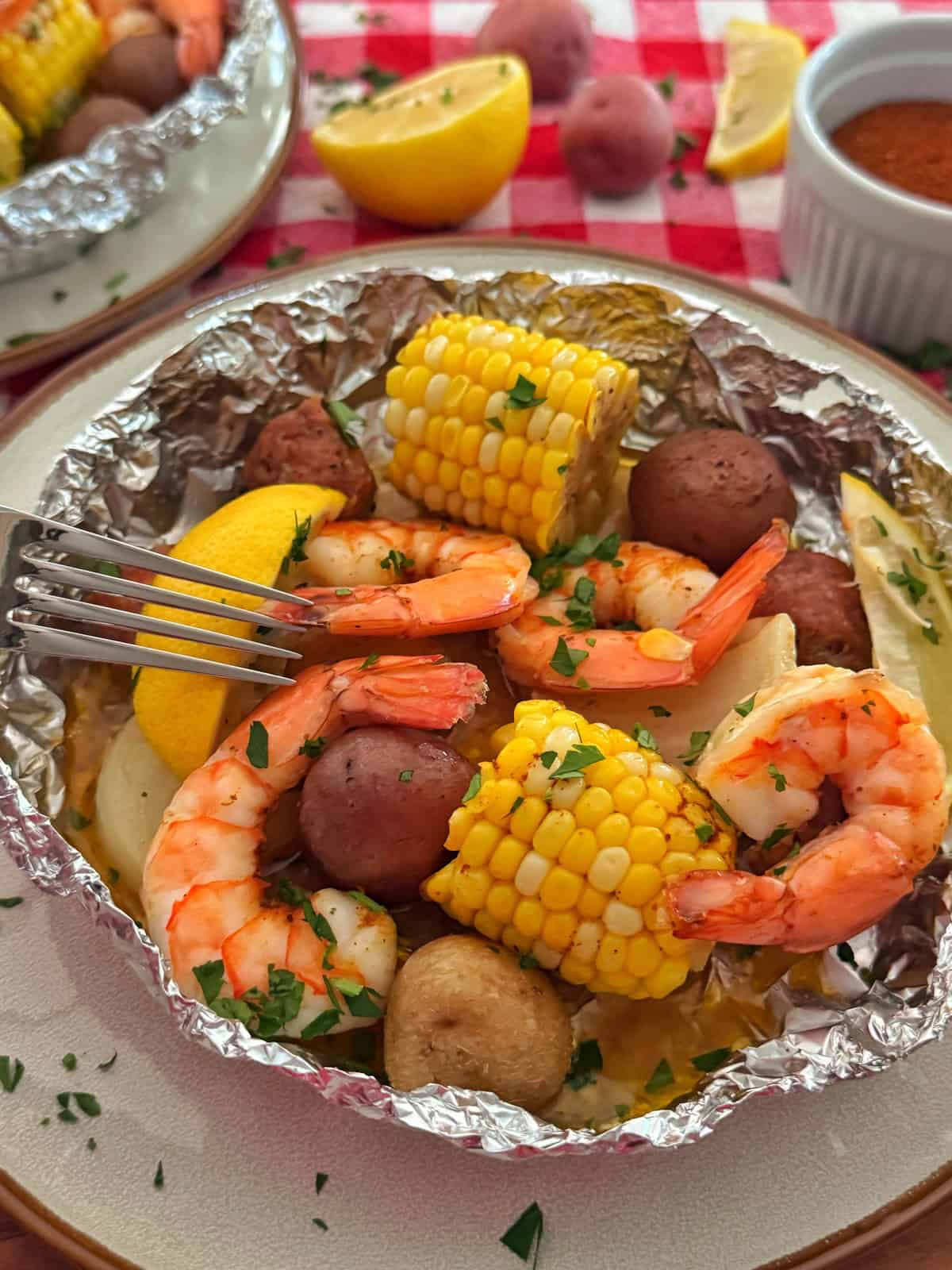 A foil packet filled with shrimp, corn on the cob, red potatoes, mushrooms, and lemon wedges, garnished with herbs. A fork rests on the food. A plate, lemon, and sauce are in the background on a red checkered tablecloth.
