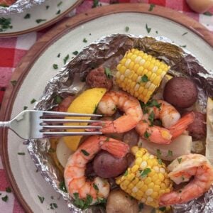 A shrimp boil foil packet filled with shrimp, corn on the cob, red potatoes, and sausage, garnished with chopped parsley and a lemon wedge, sits on a plate atop a red and white checkered tablecloth. A fork rests on the food.