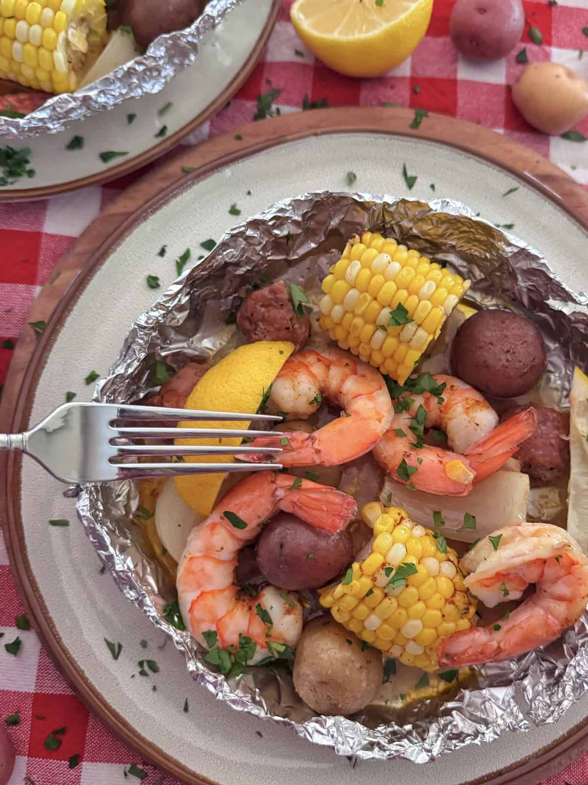 A shrimp boil foil packet filled with shrimp, corn on the cob, red potatoes, and sausage, garnished with chopped parsley and a lemon wedge, sits on a plate atop a red and white checkered tablecloth. A fork rests on the food.