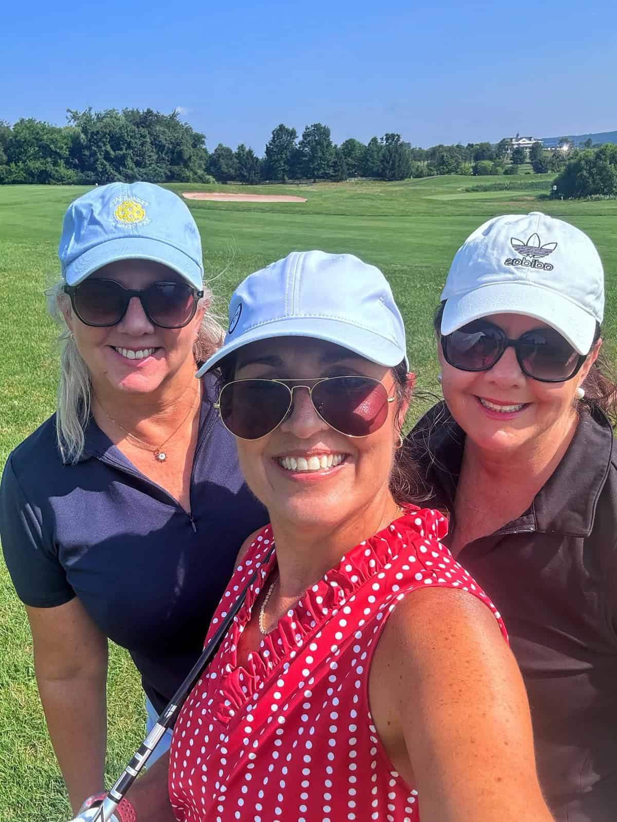 Three women wearing sunglasses and hats smile for a selfie on a sunny golf course, with green grass and trees in the background.