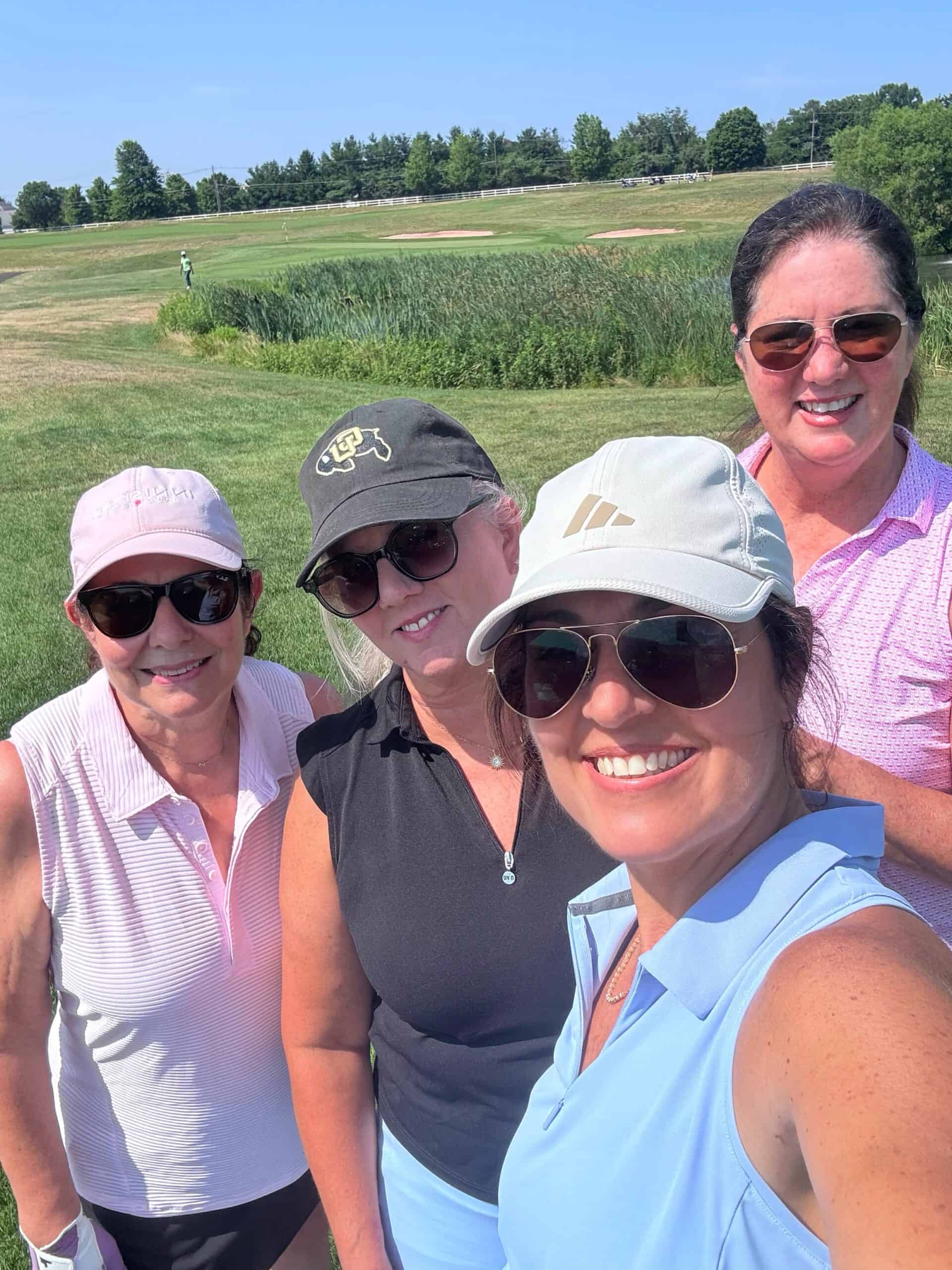 Four women wearing sunglasses and golf attire smile for a selfie on a sunny day at a golf course, with green grass, trees, and blue sky in the background.