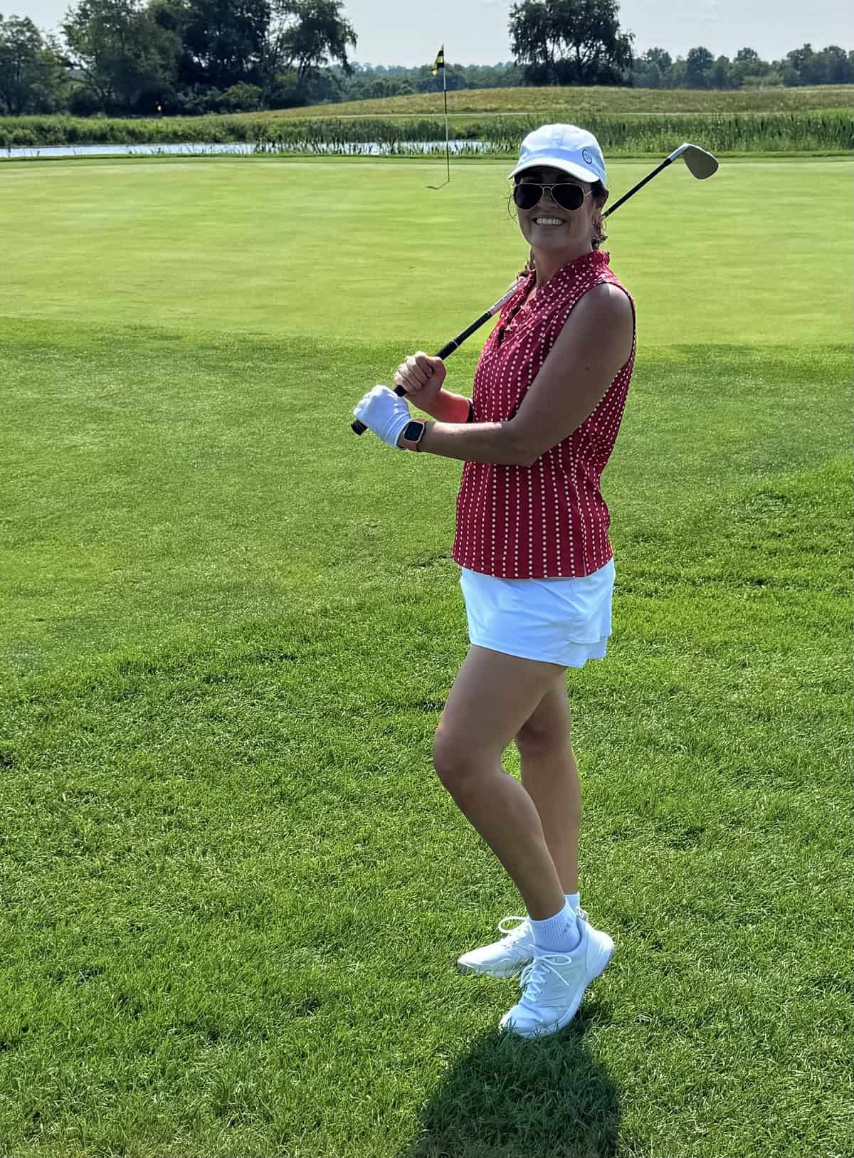 Woman in a red sleeveless top, white shorts, hat, and sunglasses stands on a golf course, smiling and holding a golf club over her shoulder. The flag and green are visible in the background.