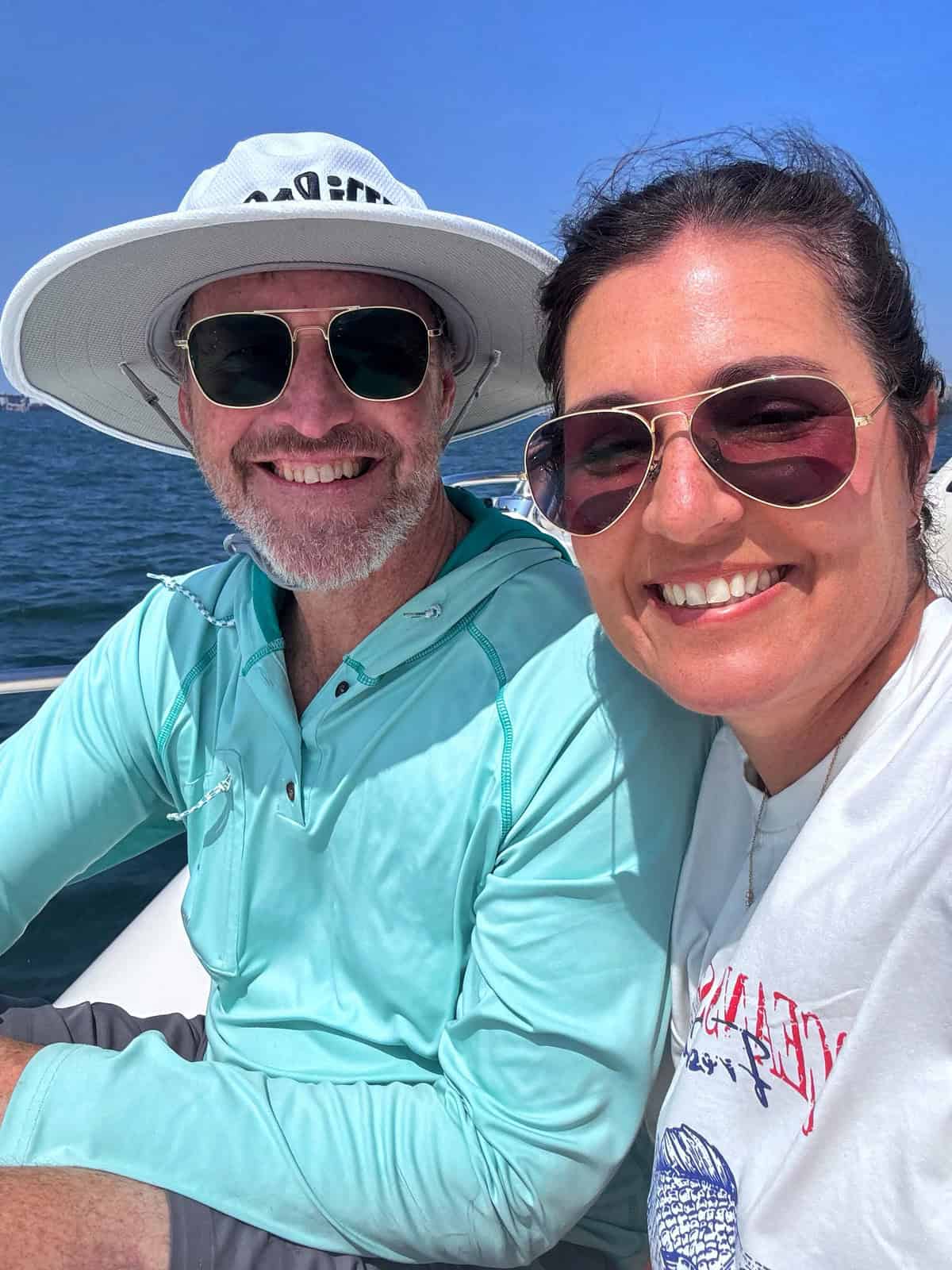 A man and woman wearing sunglasses and smiling sit together on a boat, with blue water and sky in the background. The man wears a wide hat and a light blue shirt; the woman wears a white shirt.
