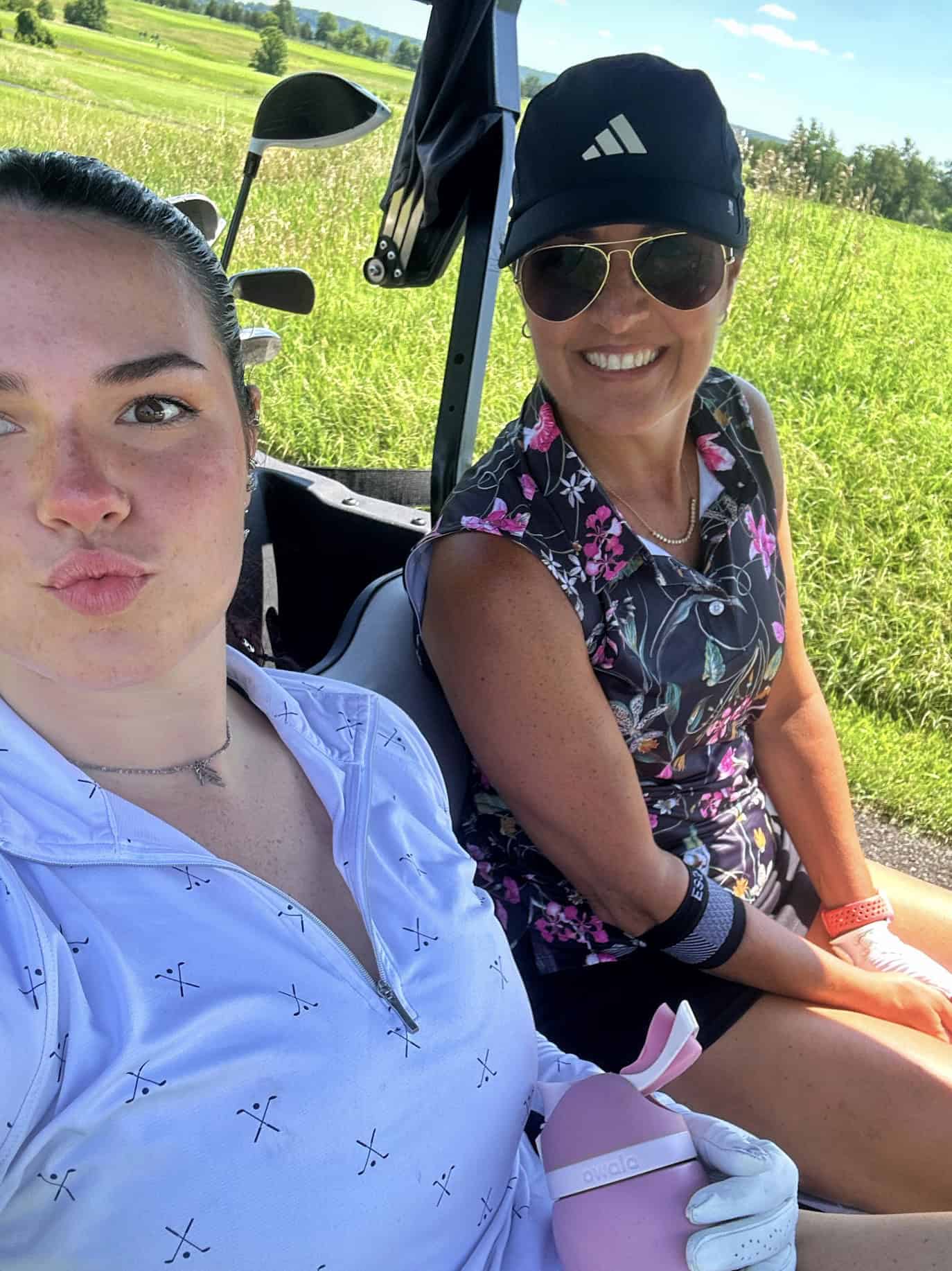 Two women sit in a golf cart on a sunny day. One takes a selfie, making a pouty face, while the other smiles at the camera. Both wear golf attire and appear relaxed, with green grass and blue sky in the background.