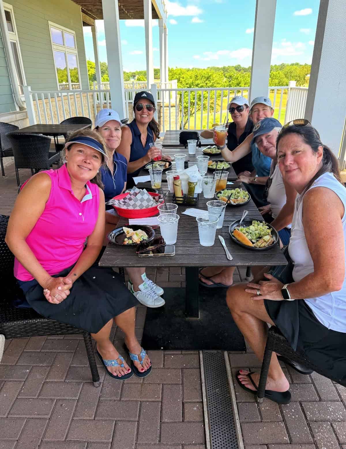 Seven women (one is gardener stacy ling) sit around a patio table enjoying lunch with salads and drinks on a sunny day, smiling at the camera with a scenic outdoor view in the background.