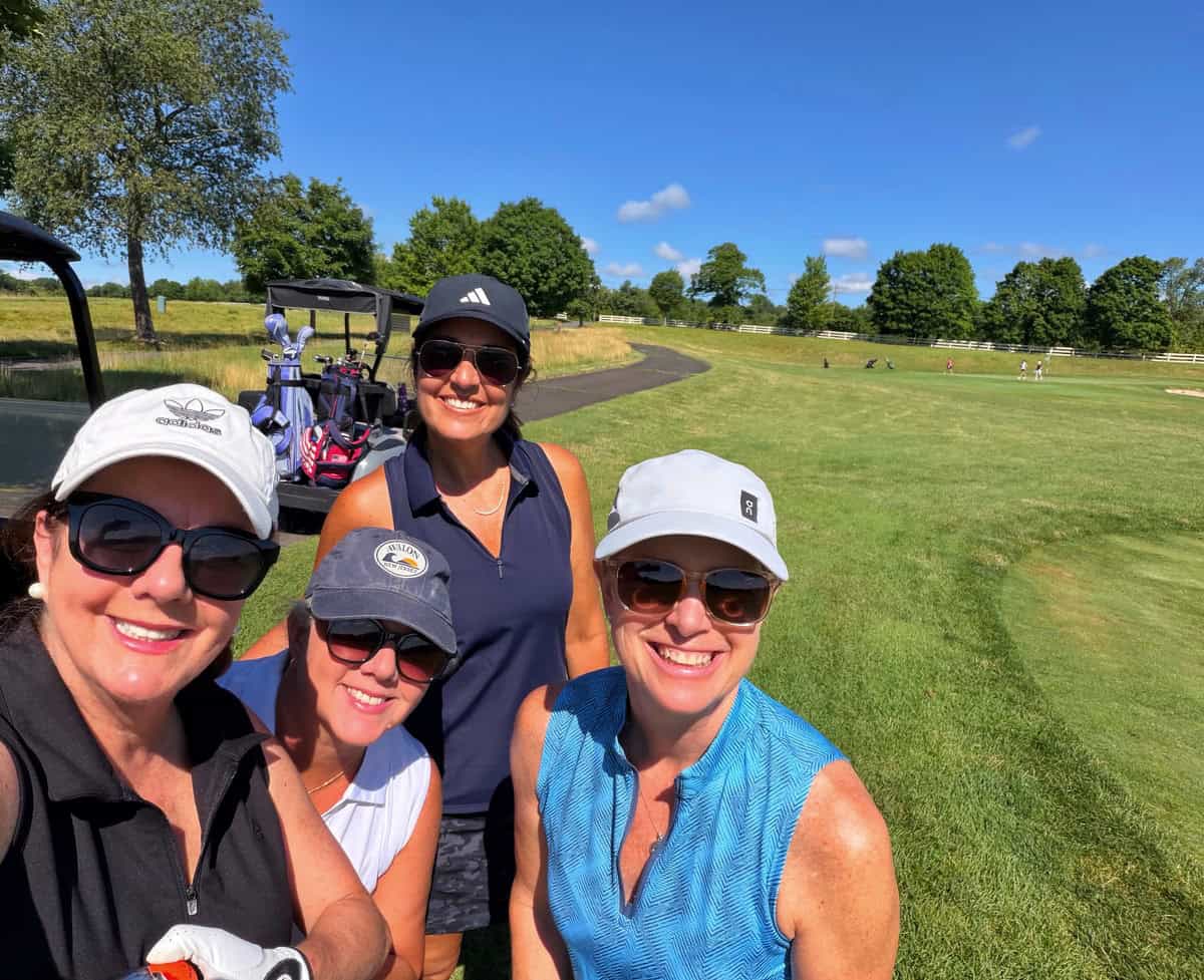 Four women in golf attire (one is stacy ling) smile for a selfie on a sunny golf course. Trees, golf bags, and a golf cart are visible in the background under a bright blue sky.