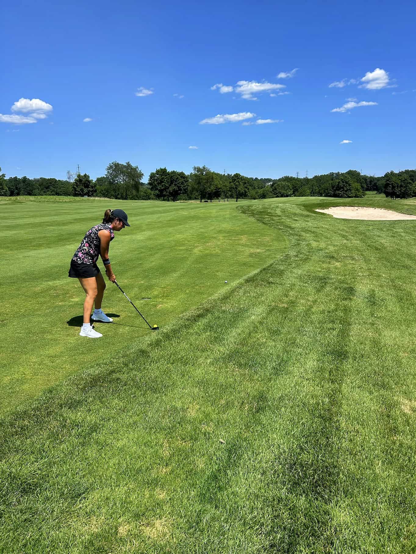 A person wearing a black cap and floral top prepares to hit a golf ball on a sunny golf course, with sand bunkers and trees in the background under a blue sky with scattered clouds.
