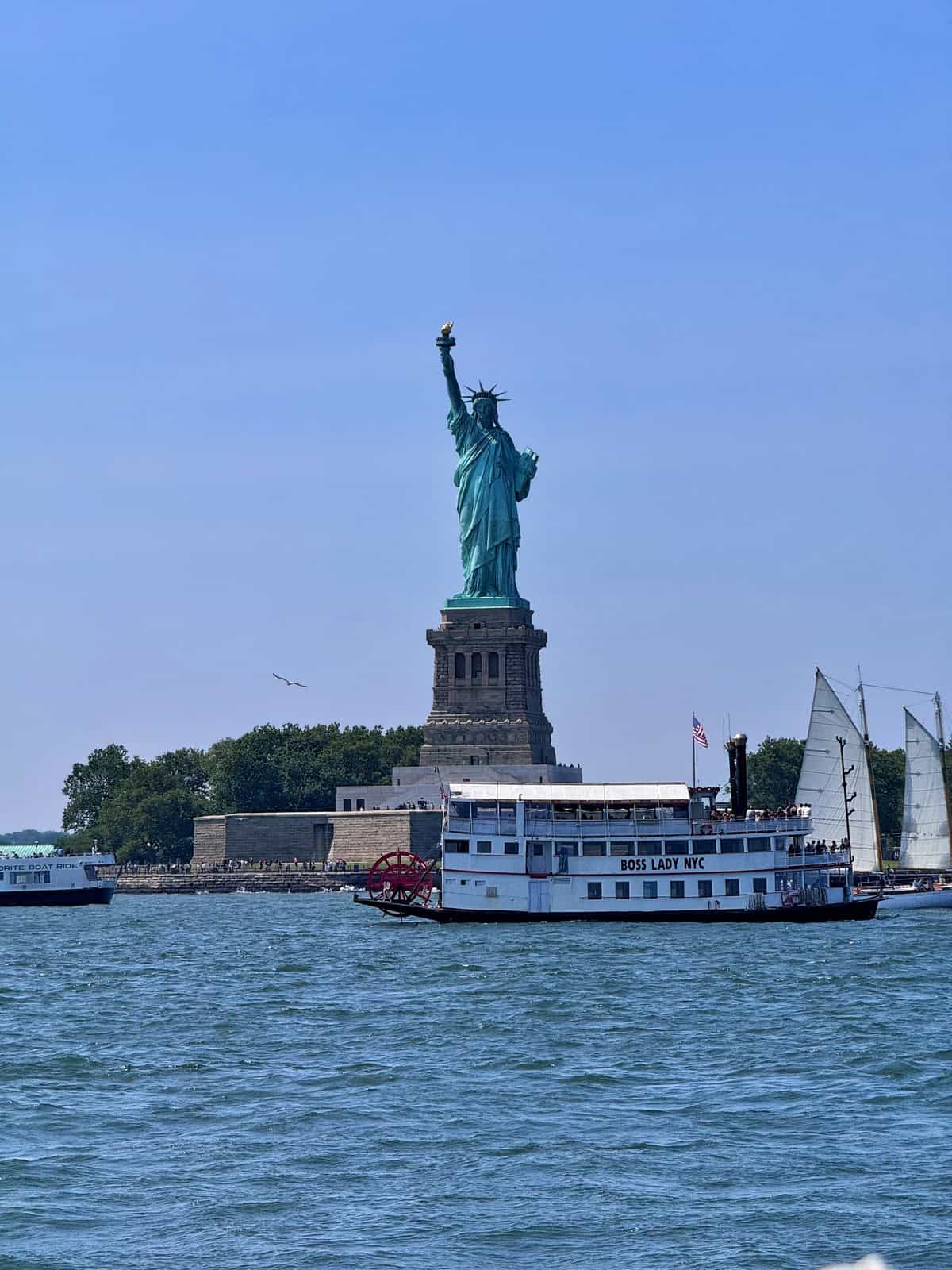 The Statue of Liberty stands on Liberty Island with two boats in the foreground, one paddleboat named "BOSS LADY NYC" and a sailboat, under a clear blue sky.