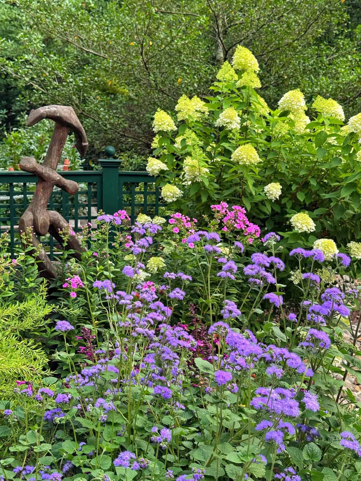 A garden with purple, pink, and pale yellow flowers, green foliage, a green fence, and a rustic wooden anchor sculpture in the background.