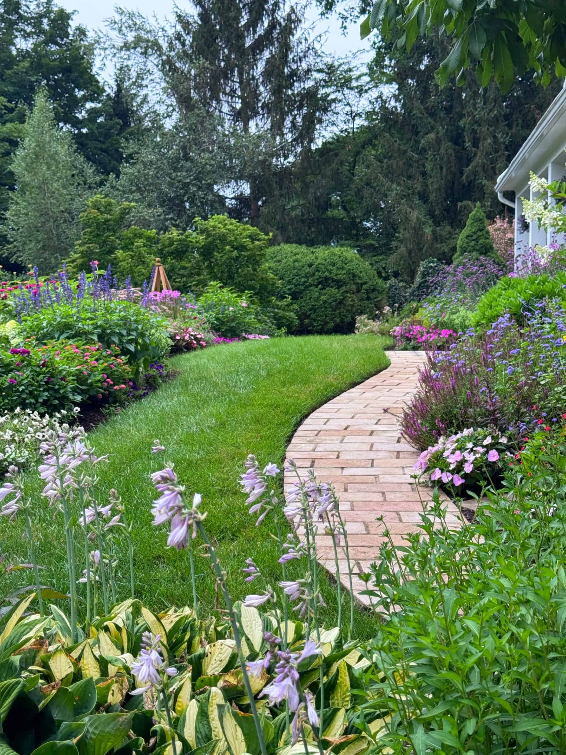 Curved brick path bordered by lush green grass and colorful flower beds, leading beside a house in a vibrant, landscaped garden with trees in the background.