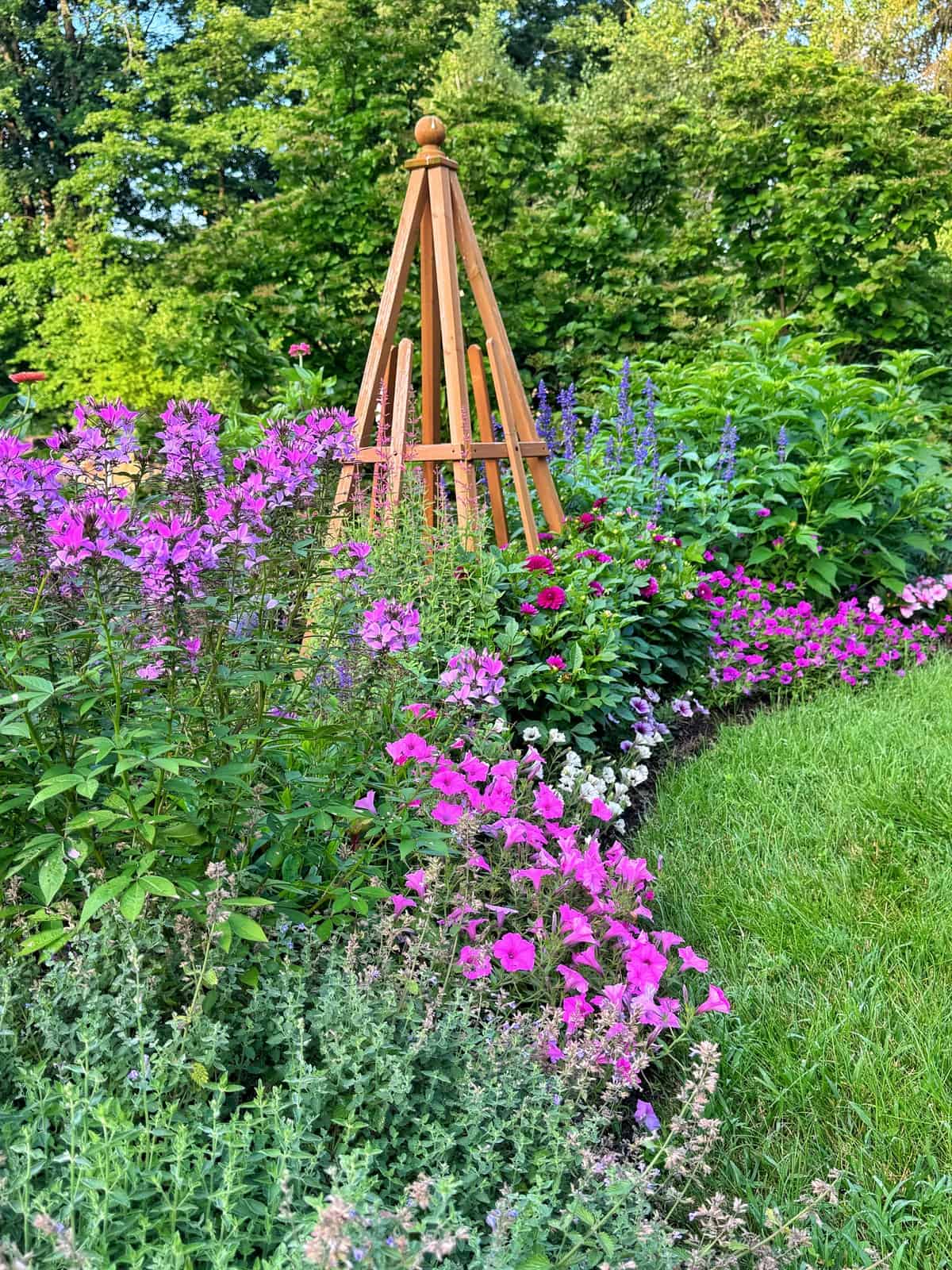 A vibrant flower garden with pink and purple blooms surrounds a wooden trellis. Green foliage and a neatly trimmed lawn complete the scene, with trees and shrubs in the background.