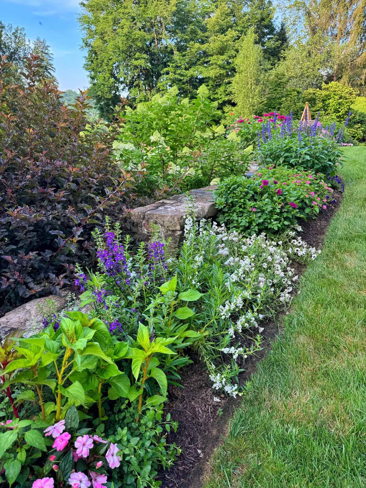 A colorful garden bed with blooming flowers in shades of pink, purple, and white, bordered by a stone wall and lush green grass, with trees and shrubs in the background under a blue sky.