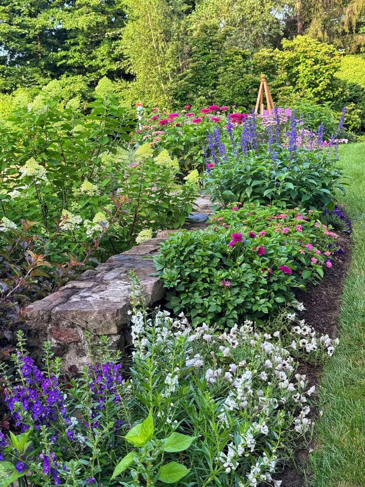 A stone garden wall is bordered by colorful flowers, including pink, purple, and white blooms, with green bushes and trees in the background on a sunny day.