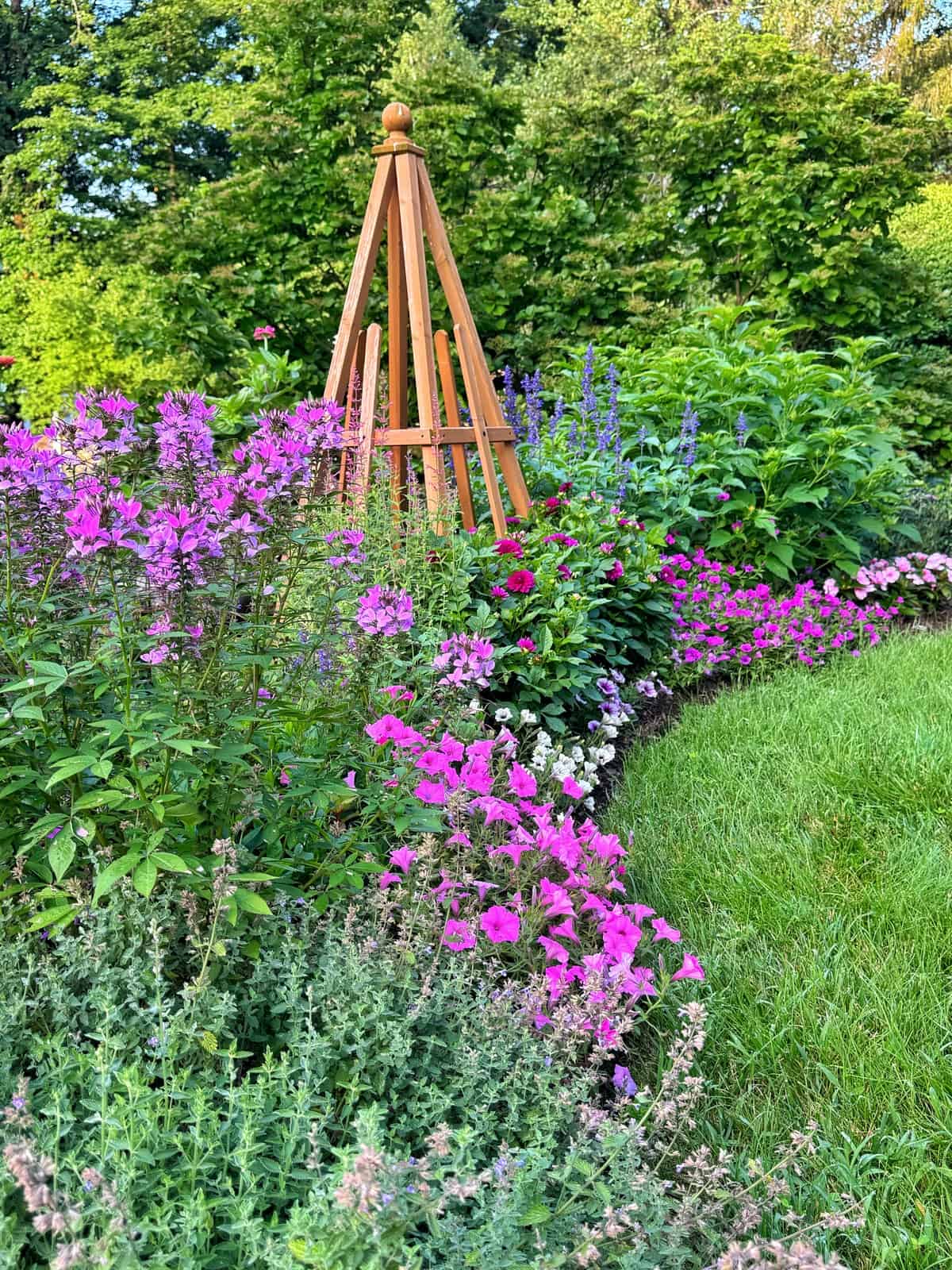 A vibrant flower garden with pink, purple, and blue blossoms surrounds a wooden garden trellis, bordered by lush green grass and trees in the background.