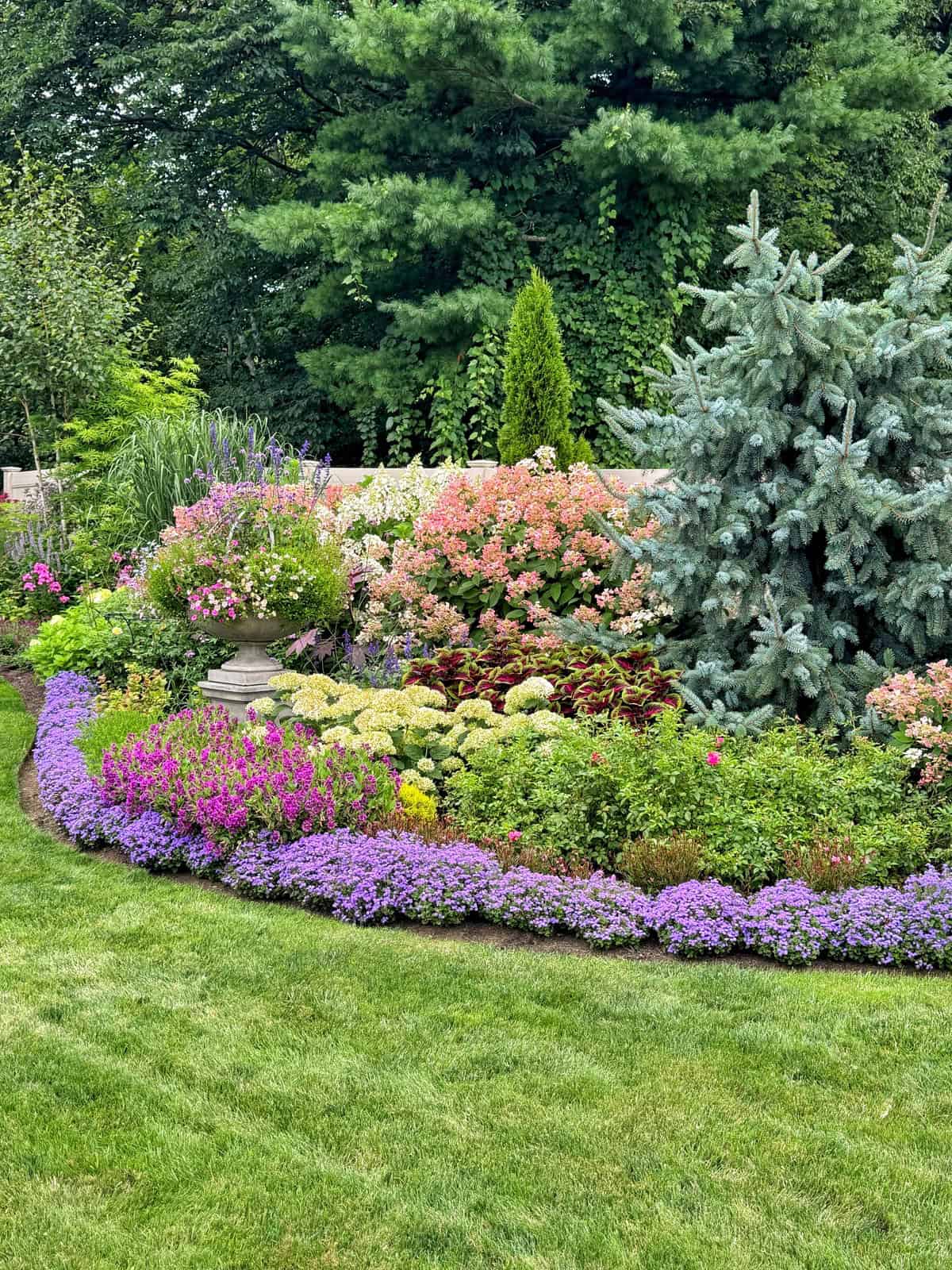 A colorful garden features vibrant purple, pink, and white flowers bordered by green grass, with lush shrubs and trees in the background.