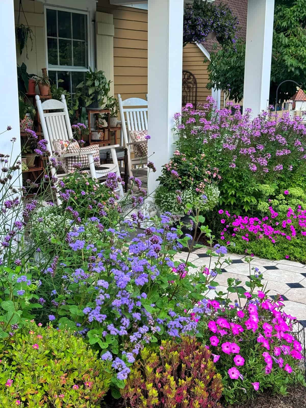 A cozy porch with white rocking chairs and potted plants overlooks a lush garden filled with vibrant pink, purple, and blue flowers on a tiled pathway.